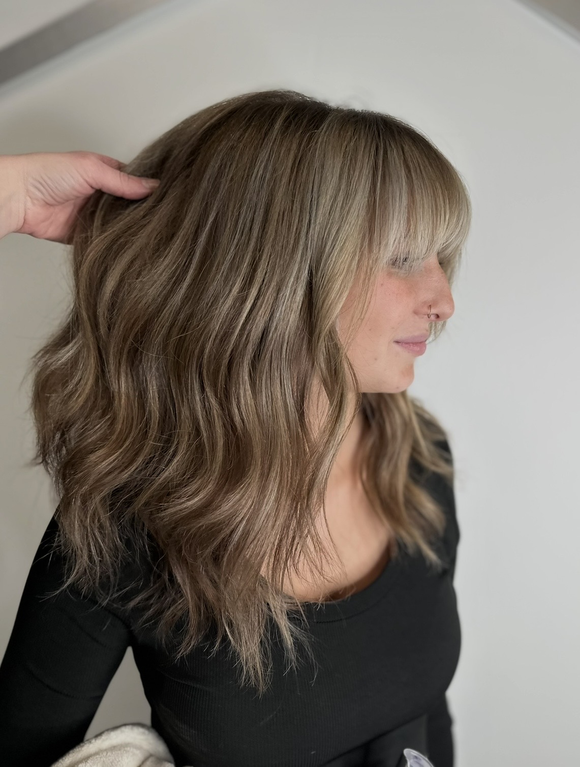 Side view of a woman with wavy, shoulder-length hair and bangs, wearing a black top, against a plain background.