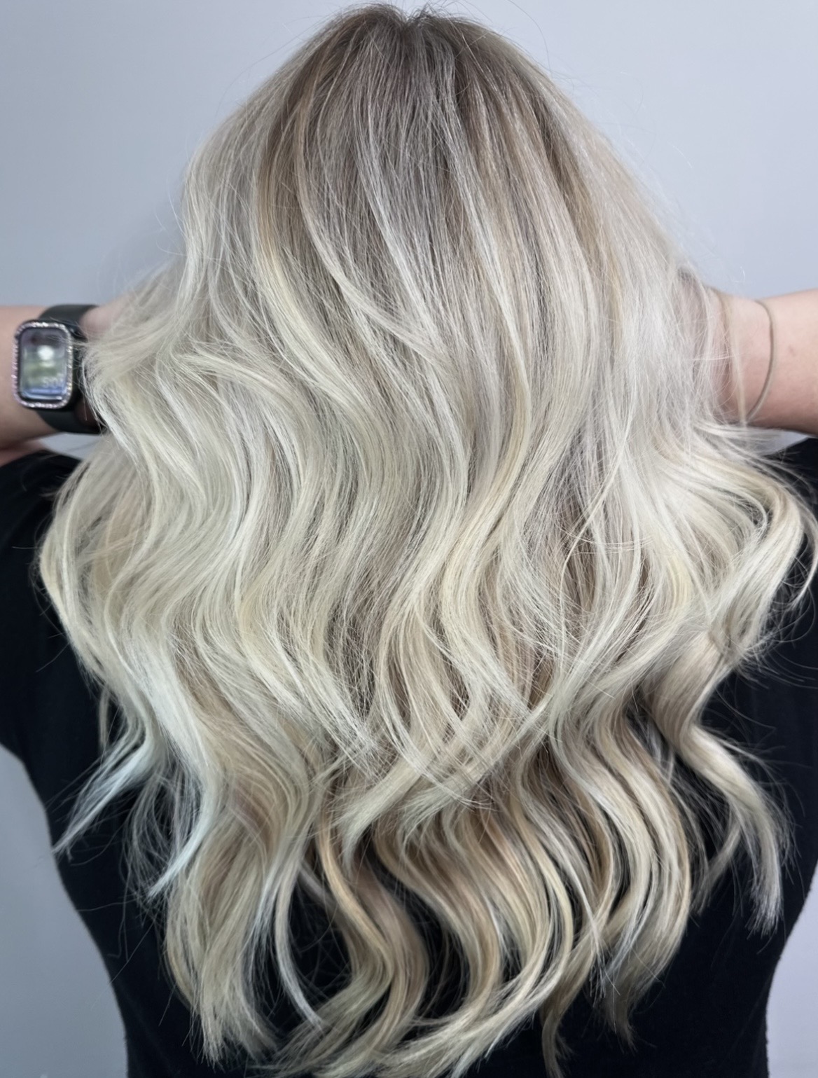 Person with long, wavy, blonde hair, viewed from the back, wearing a black top and a watch on the wrist.