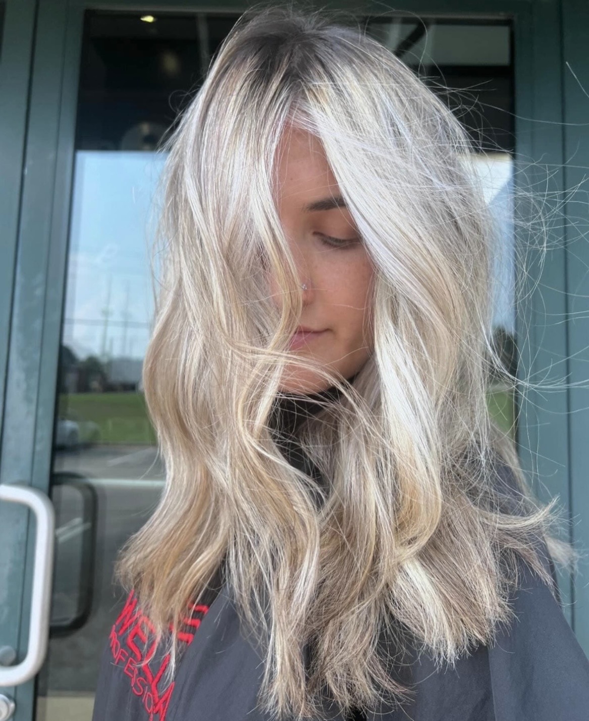 Woman with long, wavy blonde hair standing outside near a glass door, with a blurred background of a building and sky.