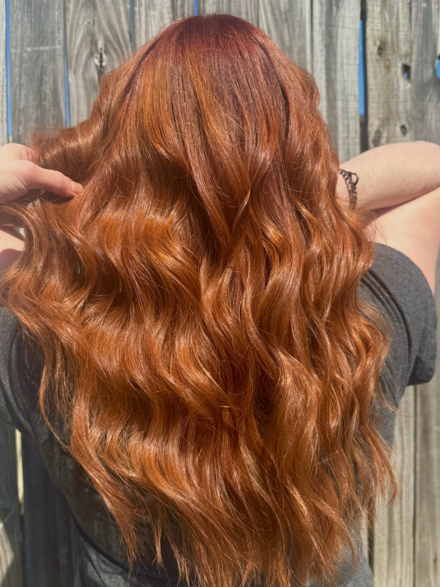 Back view of a person with long, wavy, reddish-brown hair outdoors against a wooden fence.