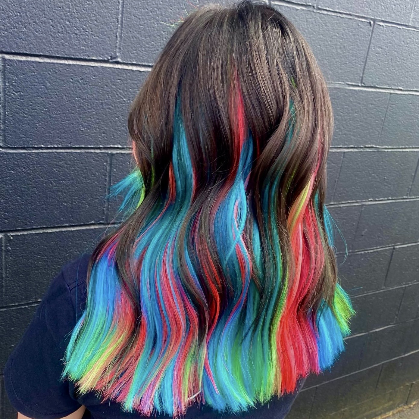 Back view of a person with shoulder-length hair dyed in rainbow colors, standing against a dark brick wall.