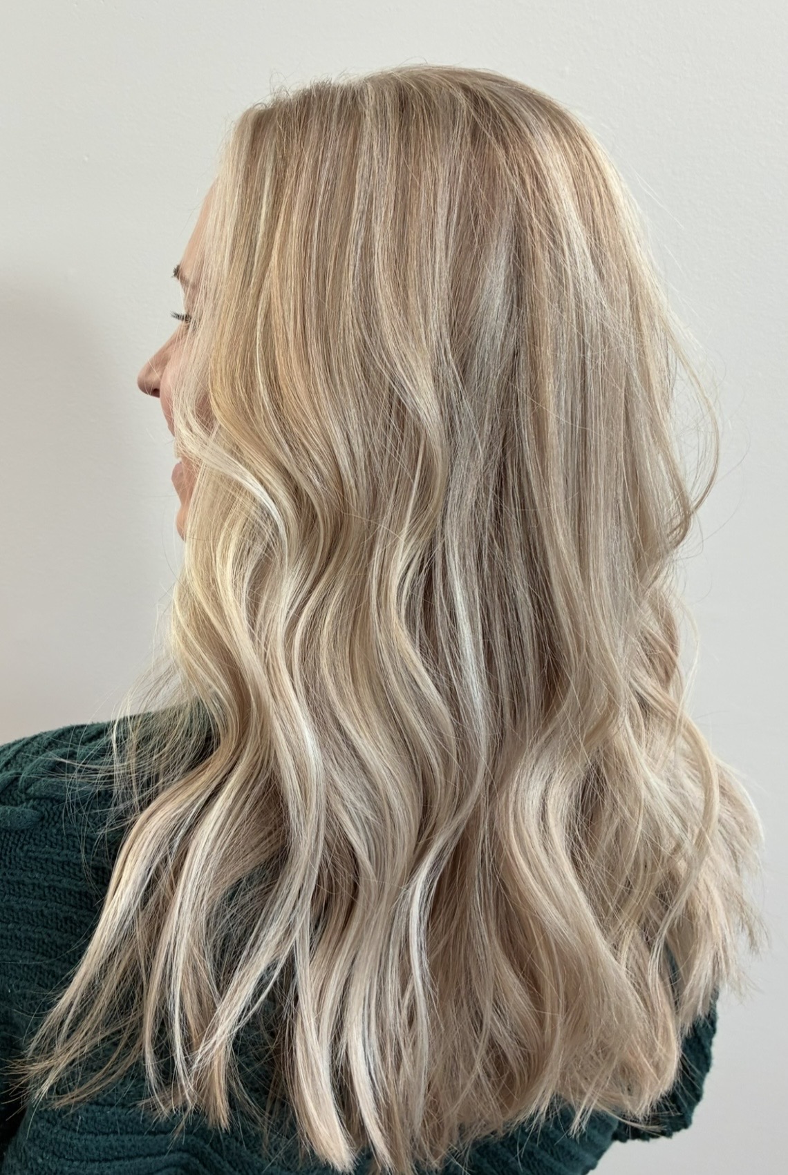 Side view of a woman with long, wavy blonde hair, wearing a dark top, against a plain background.