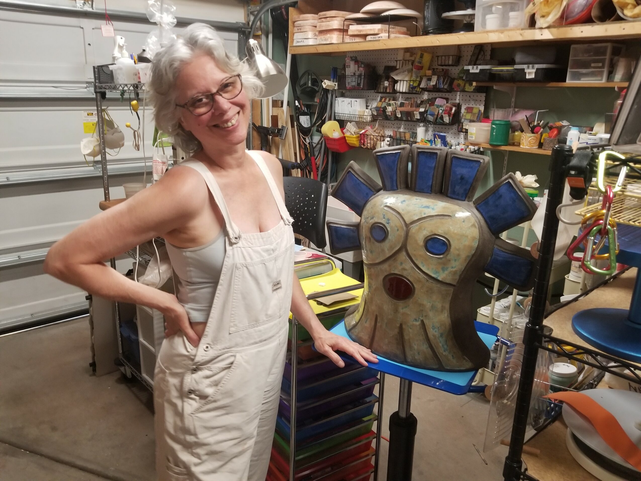 Smiling woman with gray hair and glasses in workshop, standing next to a colorful abstract sculpture with large eyes.