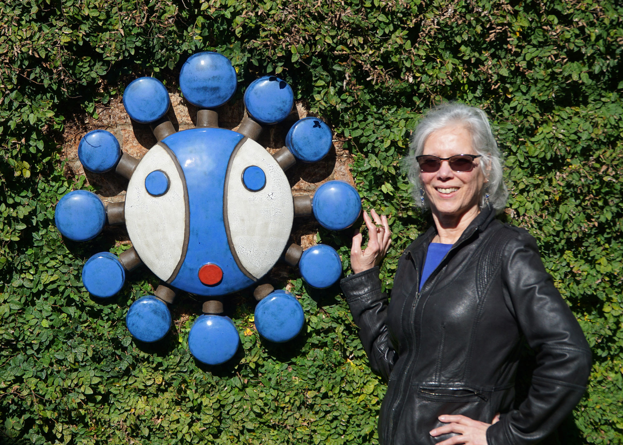 Woman with sunglasses and a leather jacket standing next to a colorful circular art piece on a green hedge.