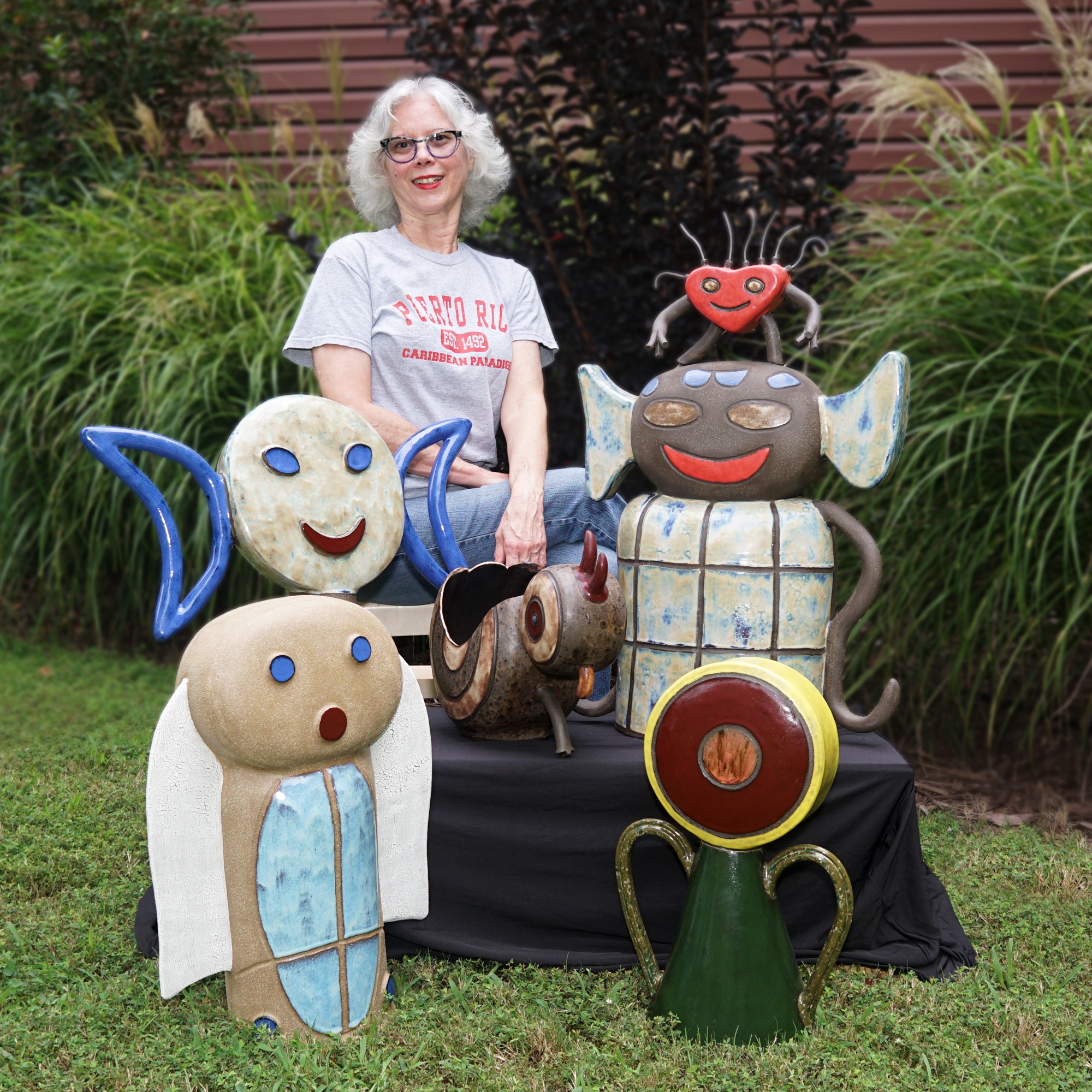 Woman with white hair and glasses standing behind colorful whimsical sculptures outdoors, surrounded by green plants.