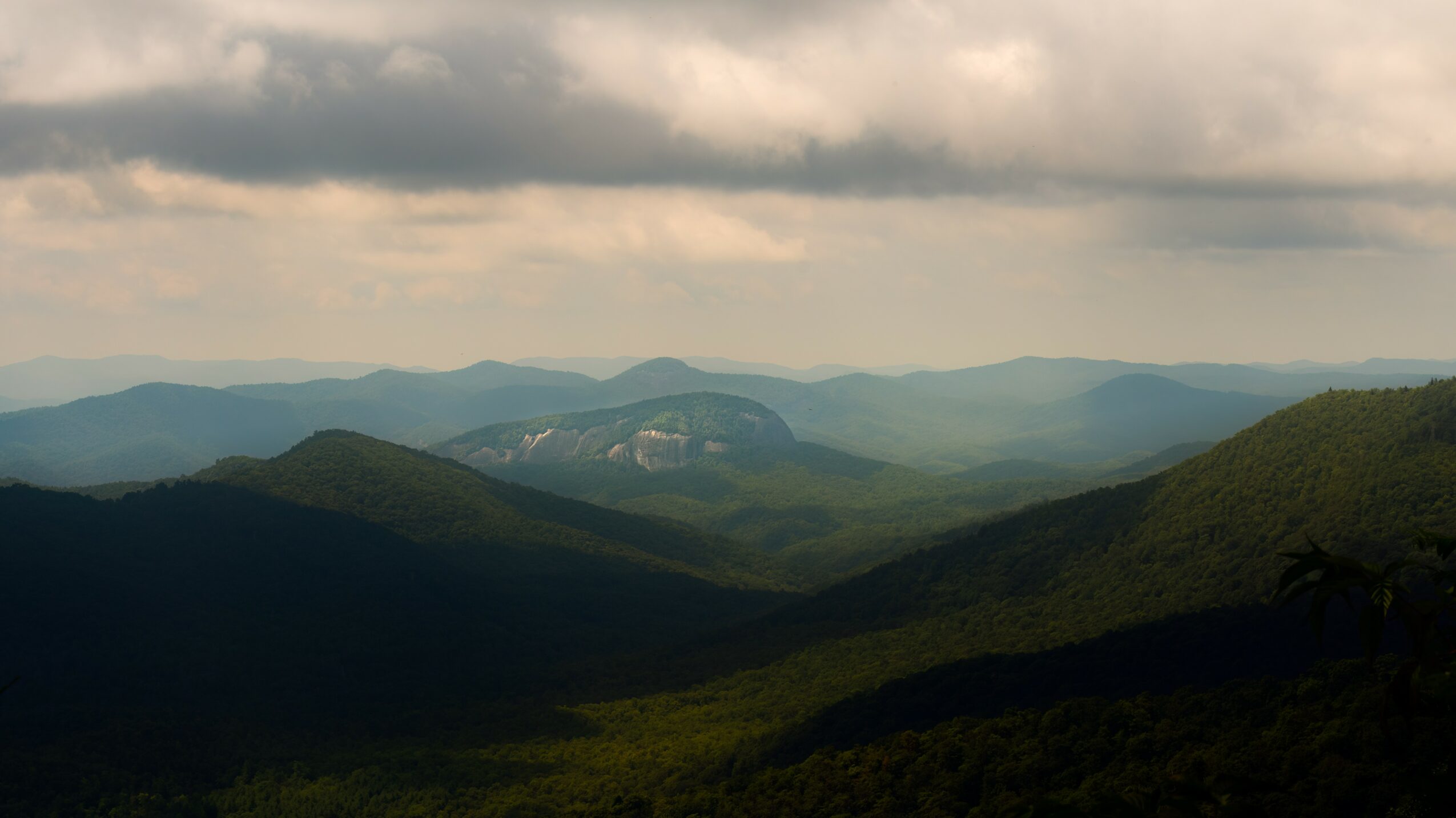 Landscape of rolling green mountains under cloudy sky, distant mountain range visible in the background.
