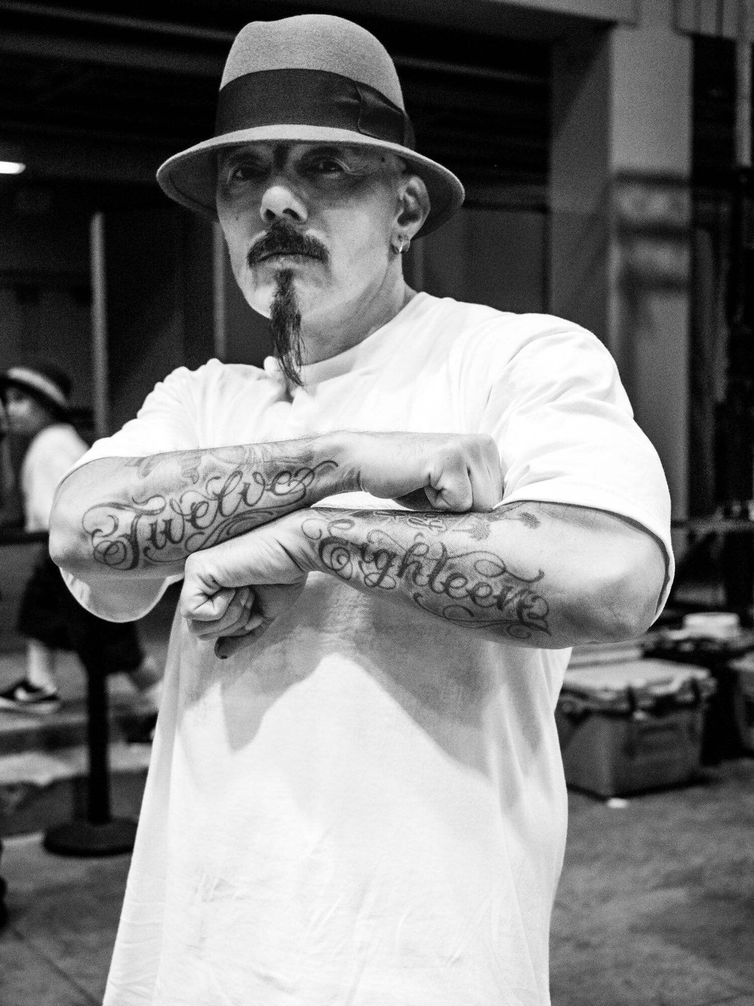 Man with a hat and tattoos crossing arms, standing outdoors in black and white.