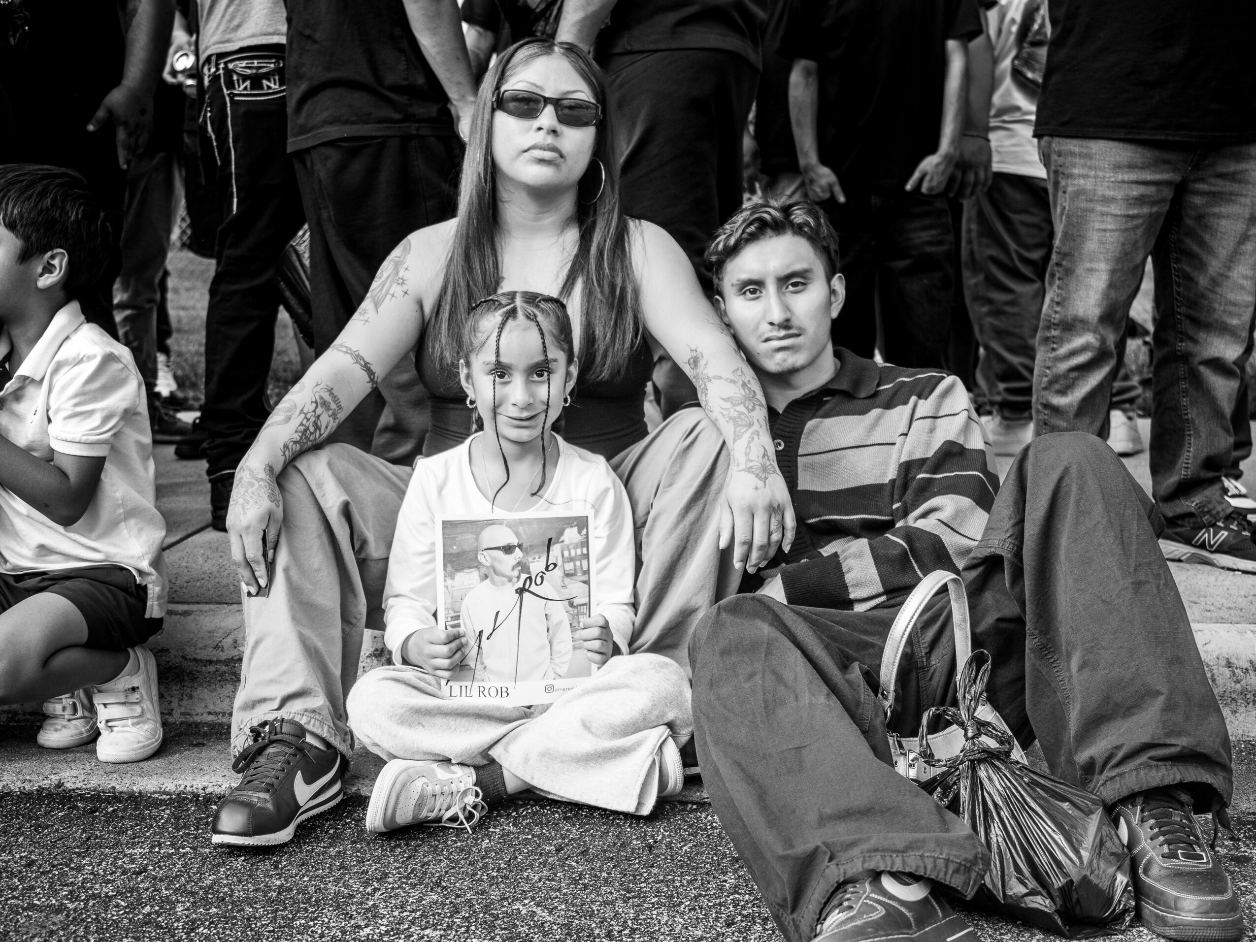 Group of people sitting and standing outdoors, including a woman, a man, and a girl sitting cross-legged, with others in the background.