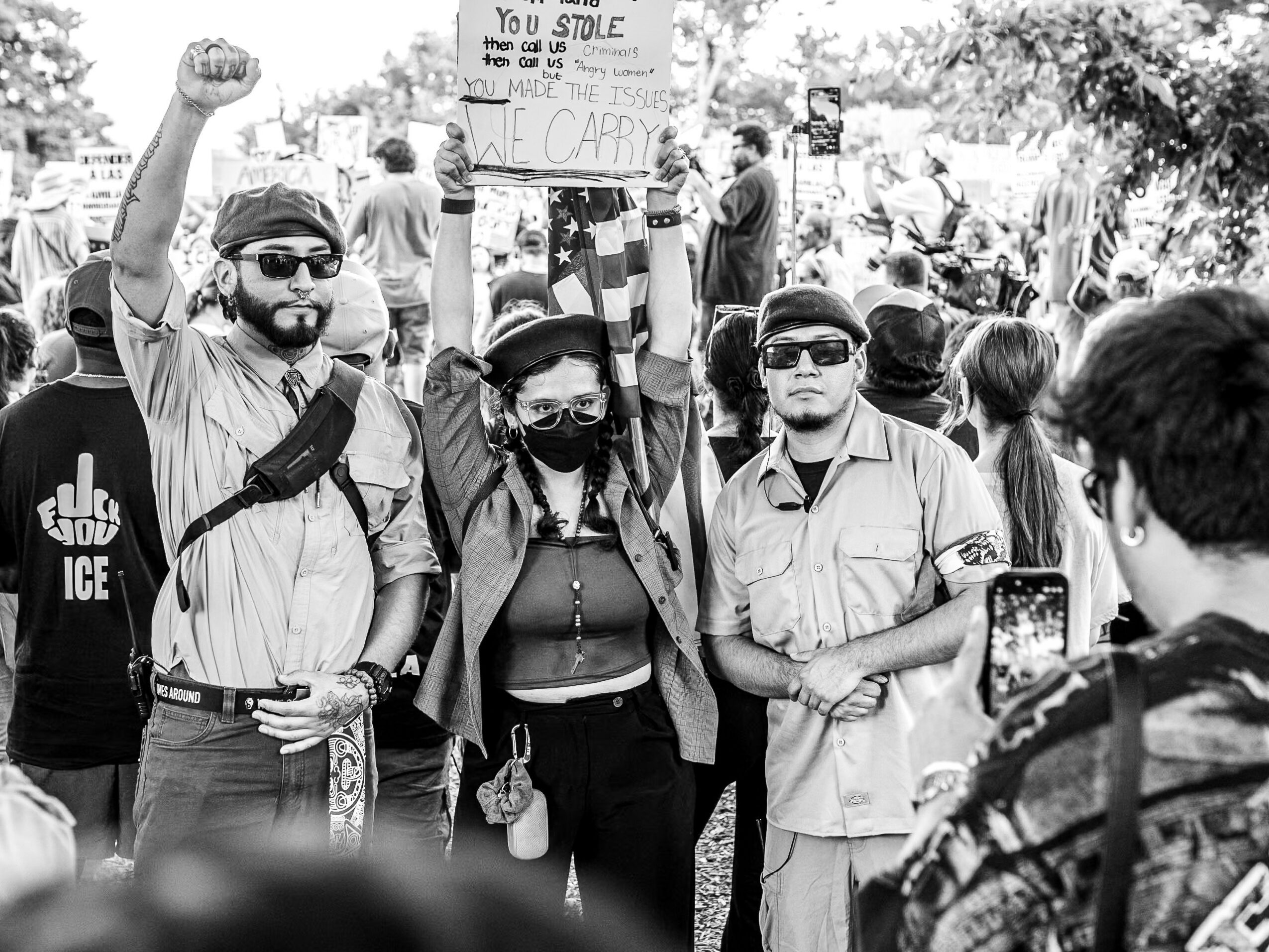 Three people holding hands and raising fists at a protest, with a crowd and protest signs in the background.