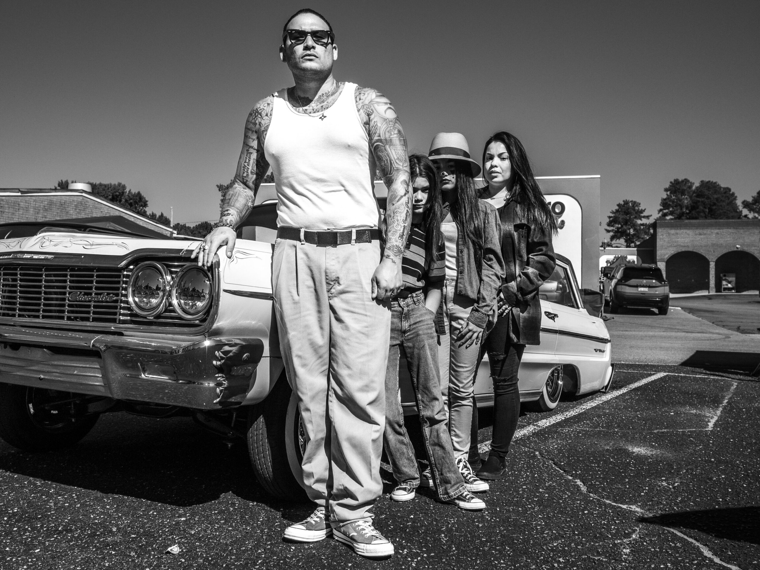 Four people standing in a parking lot next to a vintage car, with a building and trees in the background.