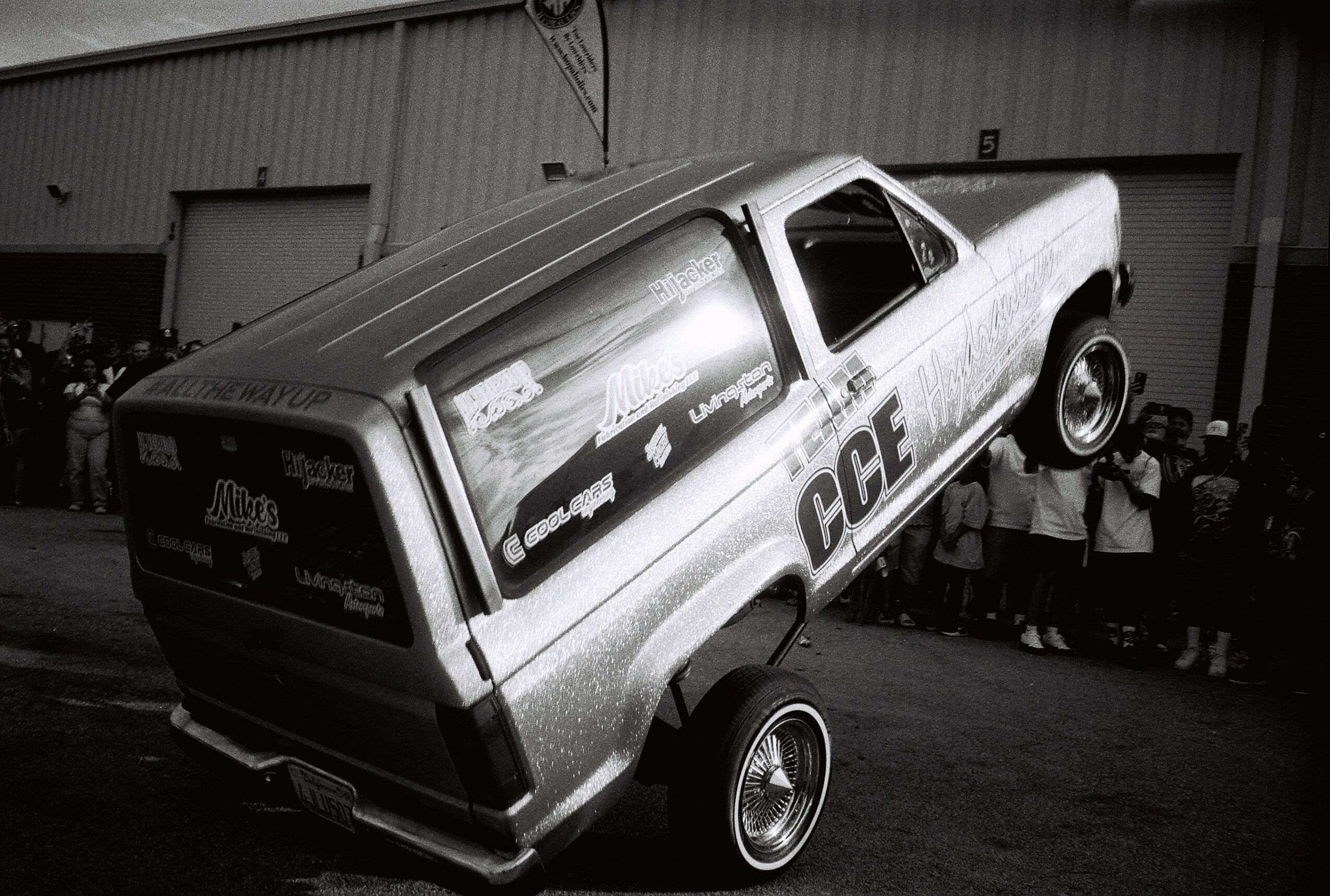 Pickup truck with front wheels lifted, parked in front of a building, with a group of people nearby, in black and white.