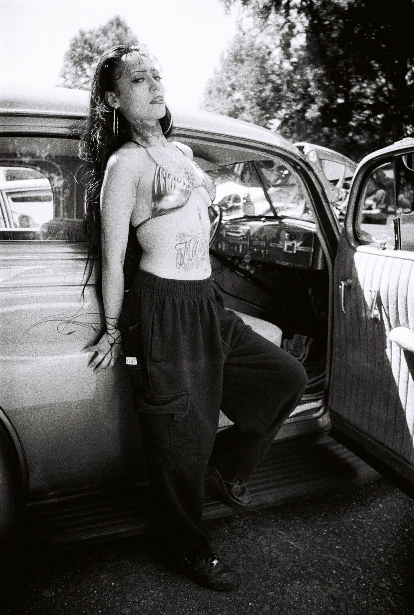 Young woman leaning against a vintage car with open door, outdoors, in black and white.