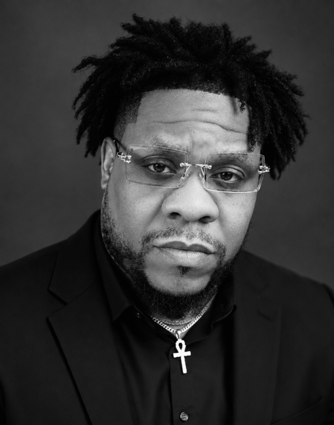 Man with glasses and an ankh necklace, wearing a dark suit, looking serious, black and white photo.