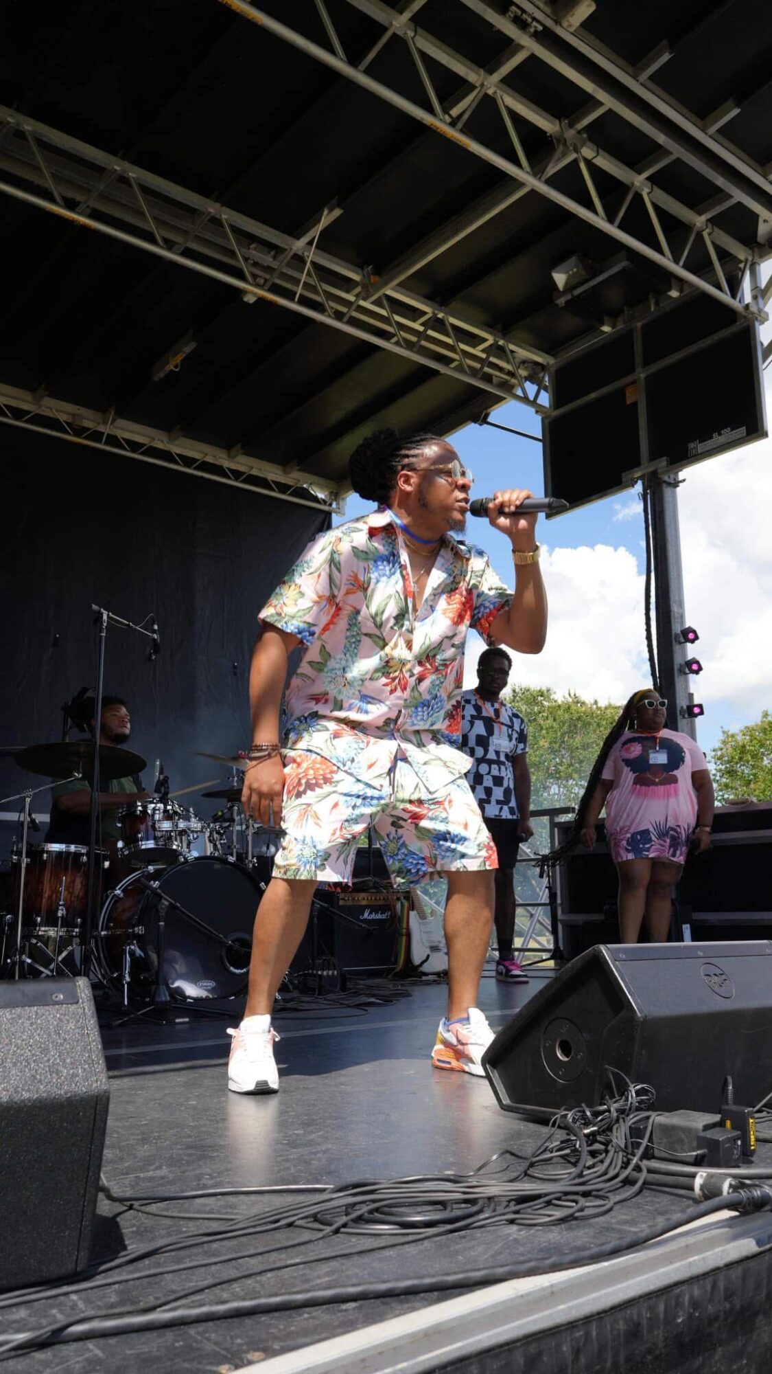 Performer singing into microphone on stage with band and audience visible, stage equipment and lighting above, outdoor setting.