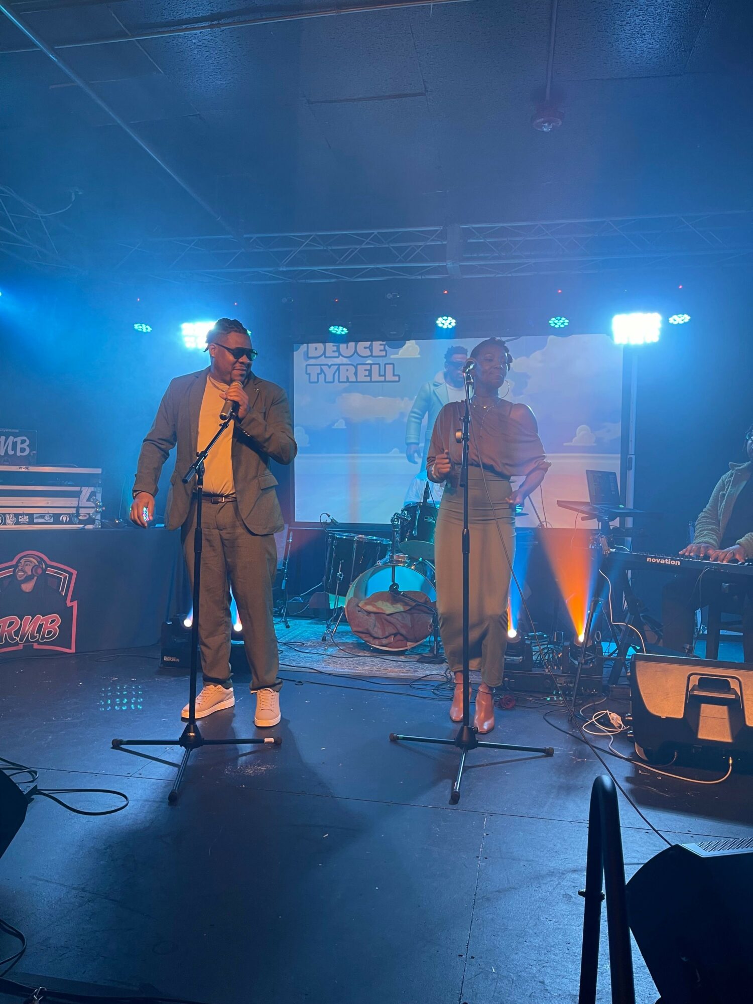 Two performers on stage with microphones, one in a beige suit and the other in a pink outfit, under blue lighting.