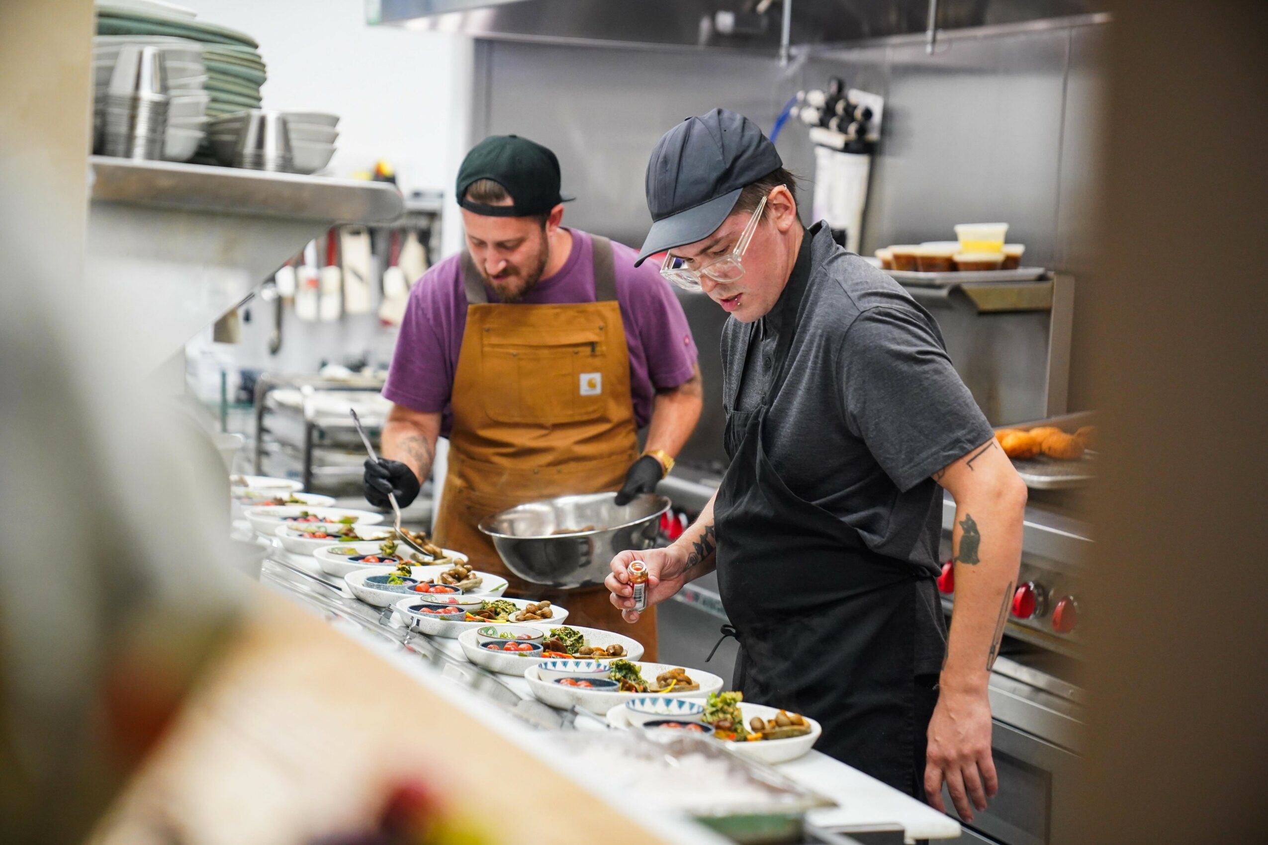 Two chefs prepare dishes in a professional kitchen, focusing on plating food on a long counter.