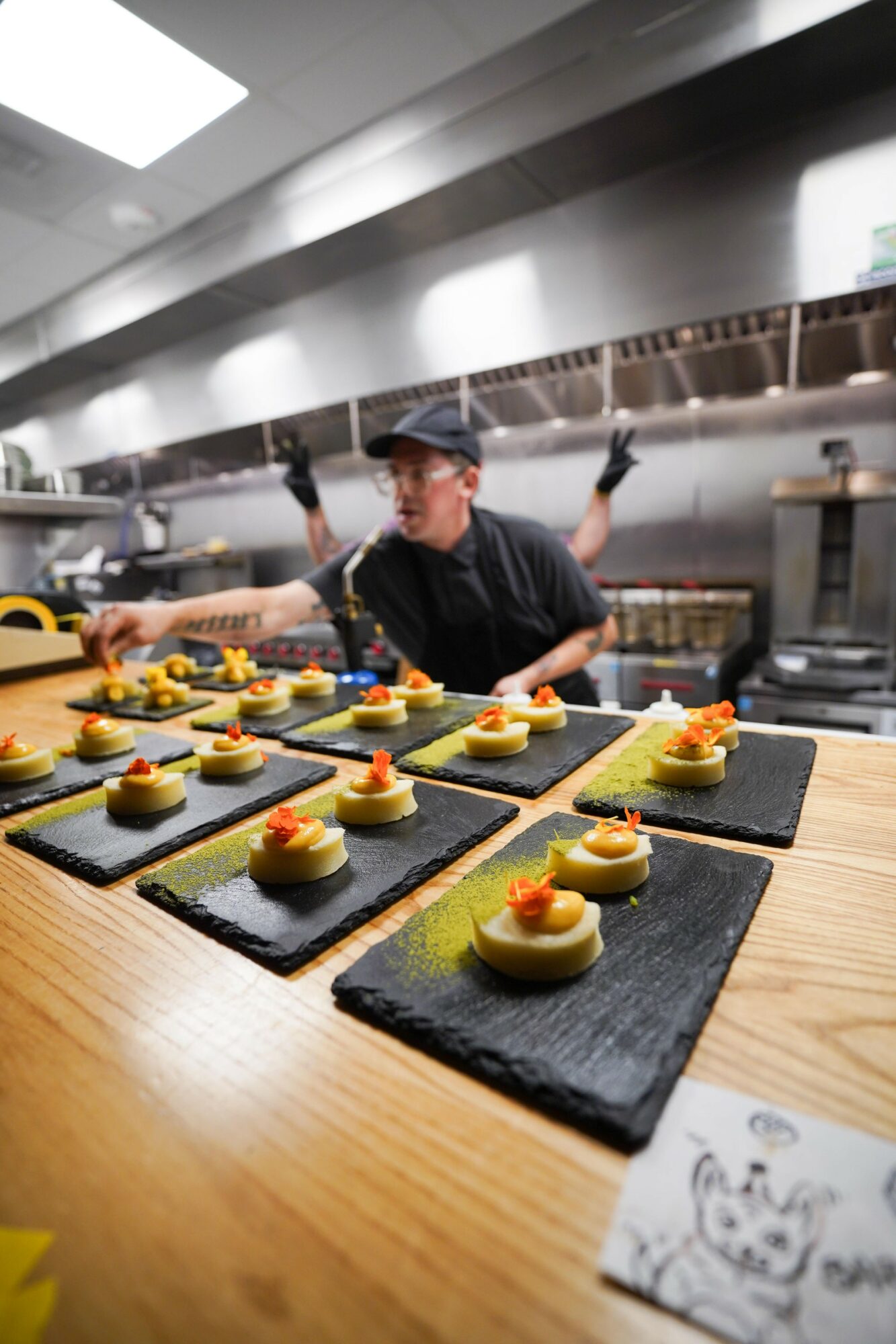 Chef preparing small dishes with yellow and red toppings on black slate plates in a professional kitchen, with stainless steel equipment in background.