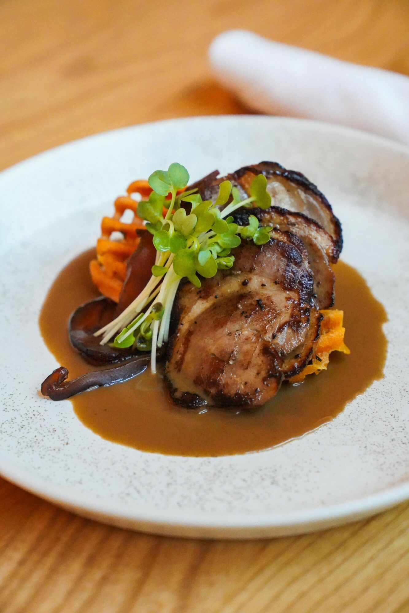 Seared meat with sauce, garnished with microgreens and vegetables on a white plate, wooden table background.
