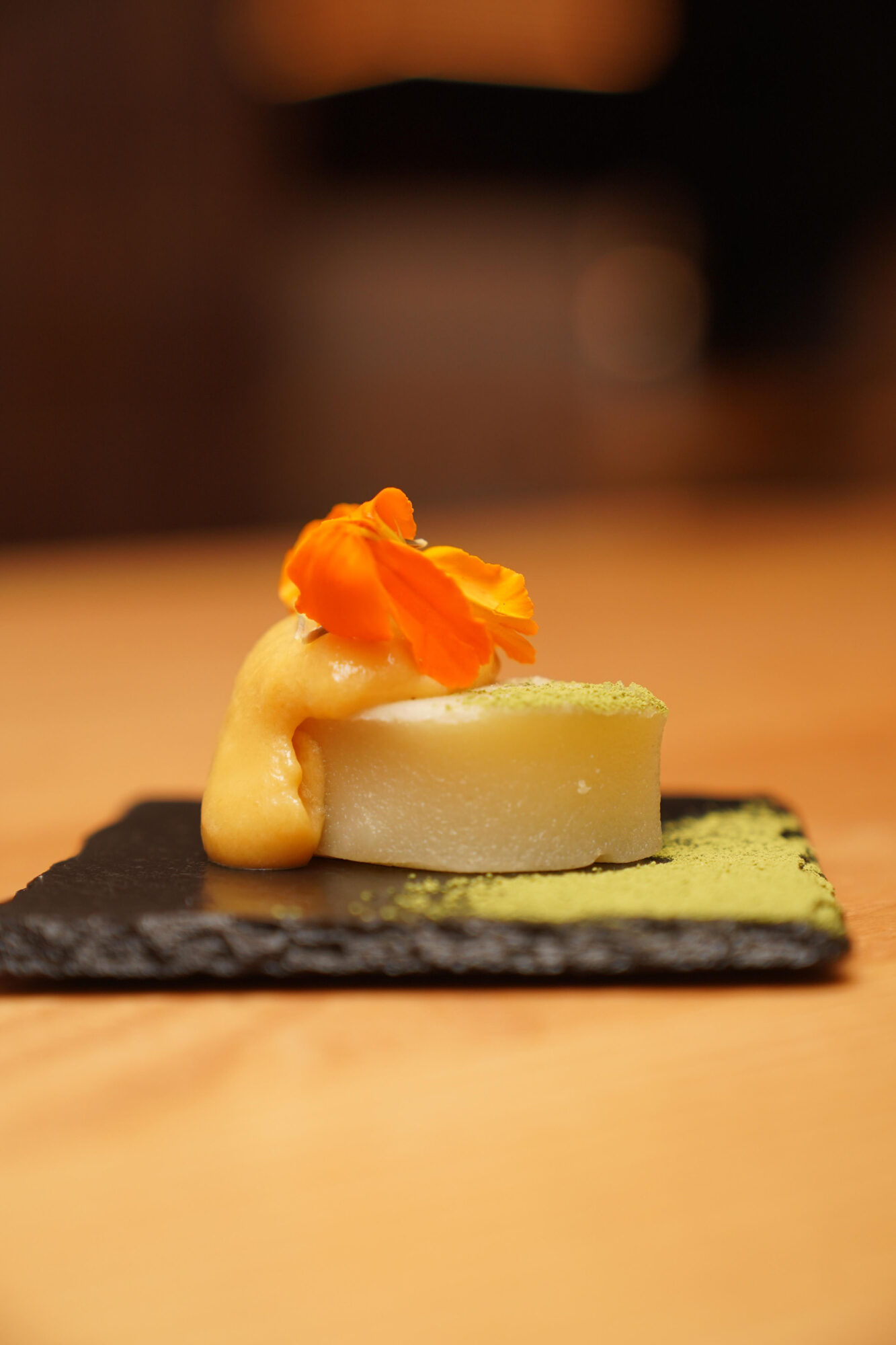 A small dish with a round, white base topped with an orange flower and a dollop of cream on a black slate plate.