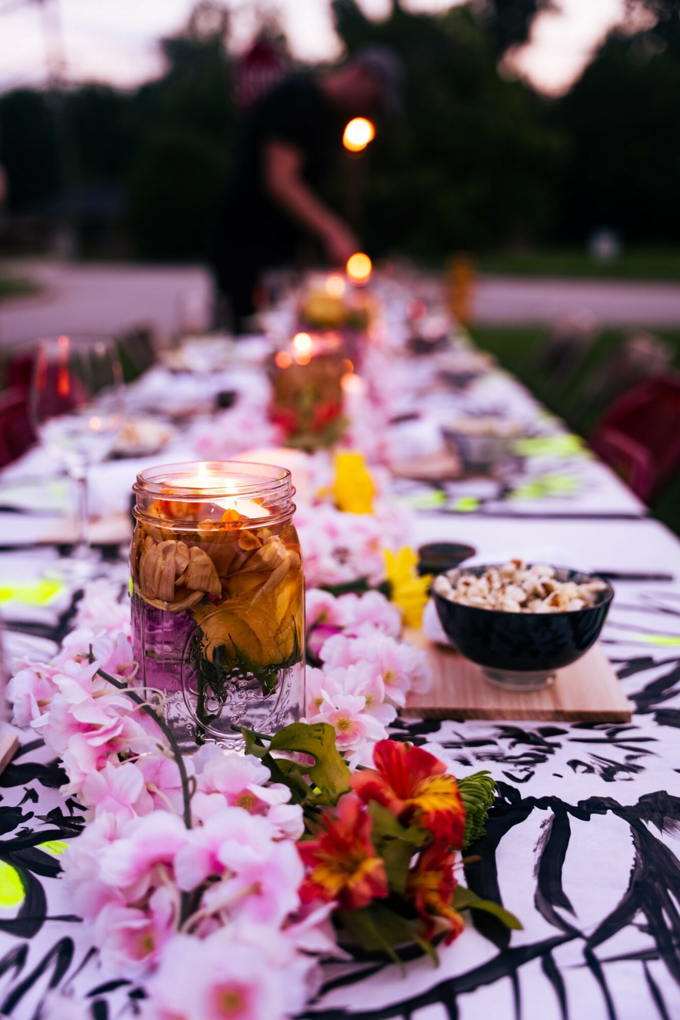 Decorated outdoor table with jars, bowls, flowers, and food, with a person in the background during evening light.