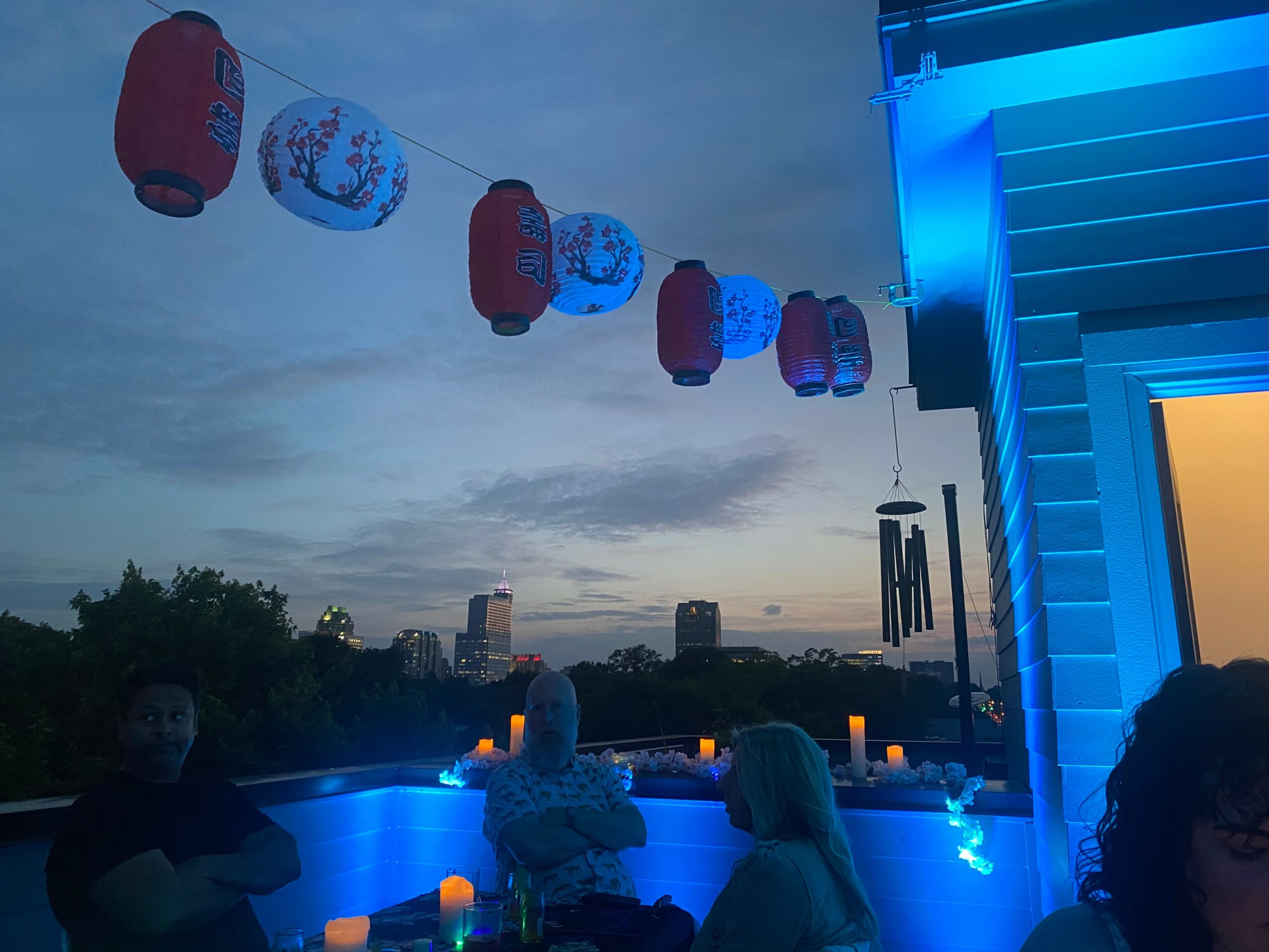 Nighttime outdoor scene with hanging lanterns, illuminated blue area, and people sitting near a building with candles.