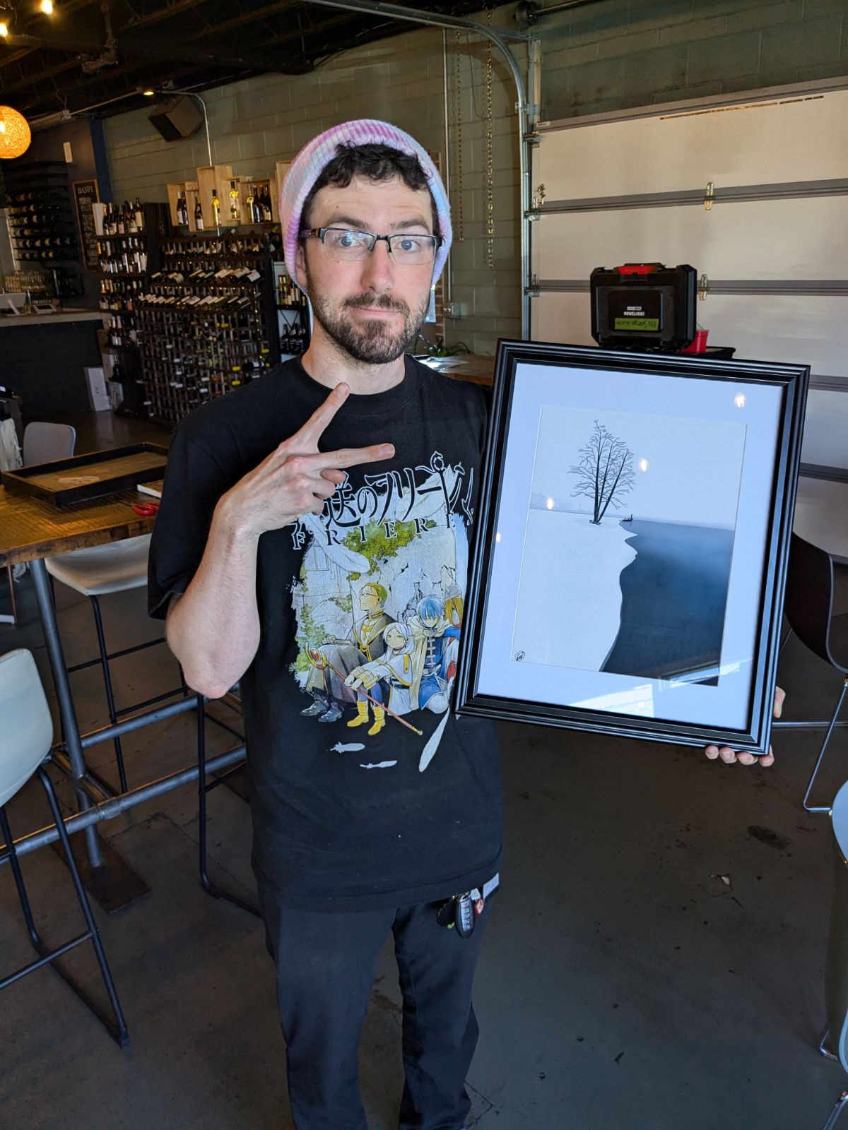 Man with glasses and a beanie making a peace sign next to a digital photo display in a cafe or bar setting.