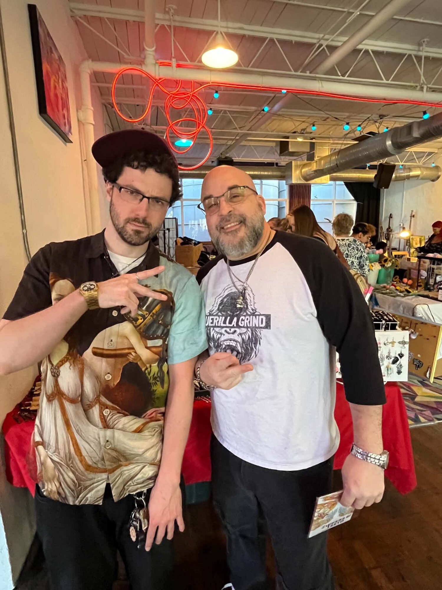 Two men standing indoors, one wearing a black hat and glasses, the other with a beard and glasses, both smiling, holding drinks.