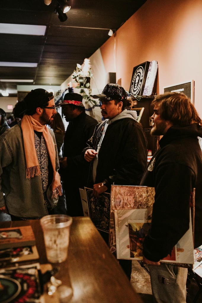Group of people browsing art and items in a gallery or store, with shelves and artwork on the wall behind them.