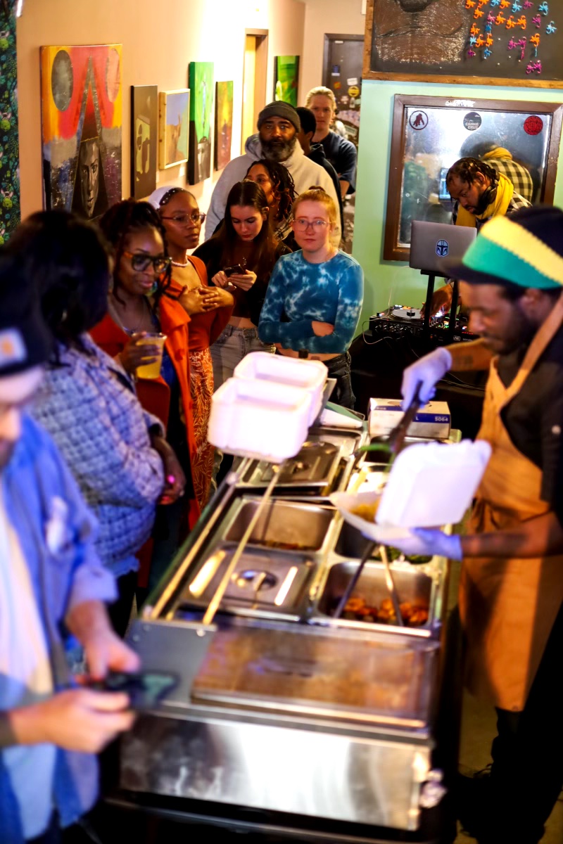 People gather around a food counter with staff serving dishes in a room decorated with artwork and a television.
