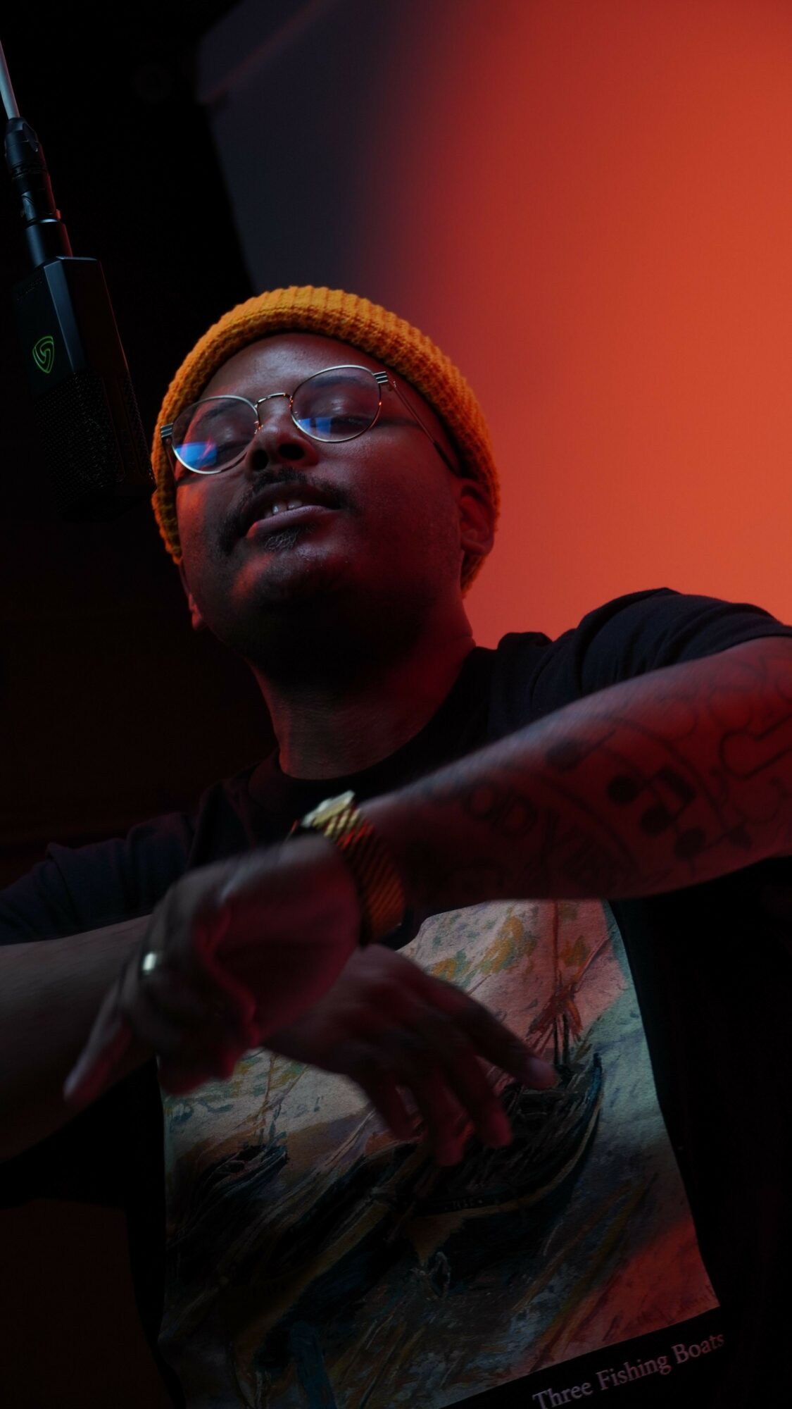 Person wearing a yellow beanie, glasses, and dark clothing, sitting at a table with a colorful background.