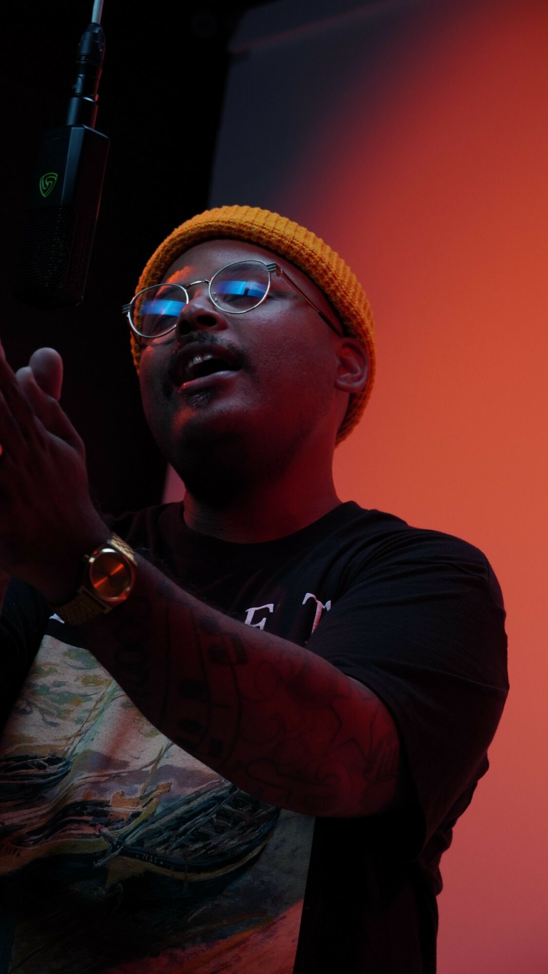 Person wearing glasses and a yellow beanie speaking into a microphone against a gradient background.