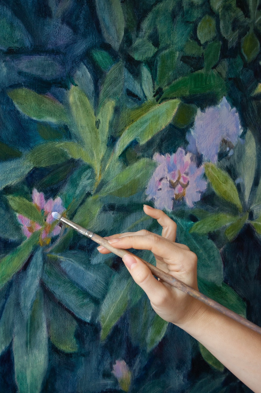 Person holding a paintbrush in front of a colorful floral painting with green leaves and pink flowers.