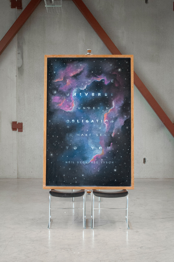 A space-themed poster with galaxy images and text, displayed on a stand with two chairs underneath, against a concrete wall.
