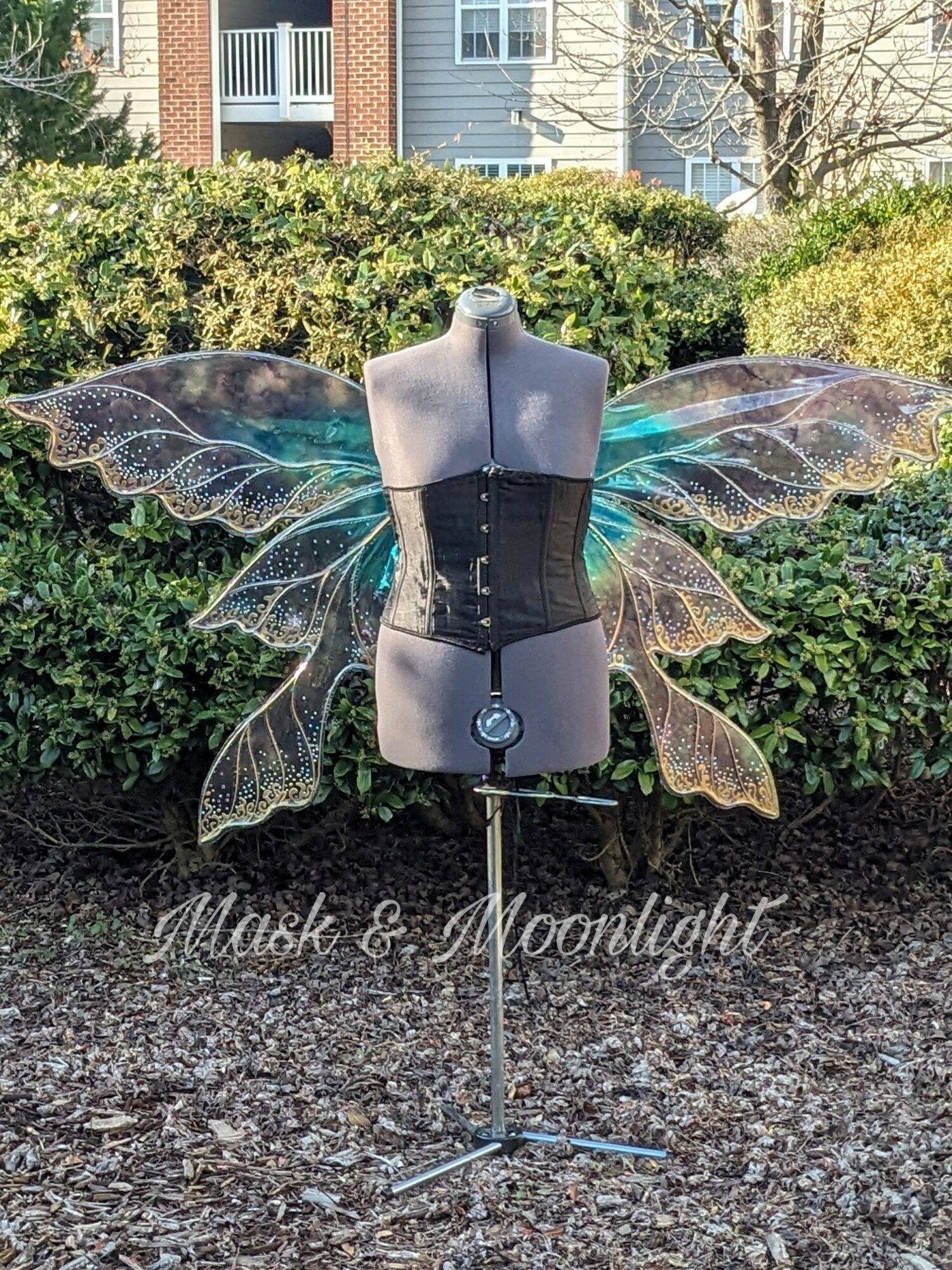 Mannequin with fairy wings, black corset, outdoors with bushes and buildings in background.