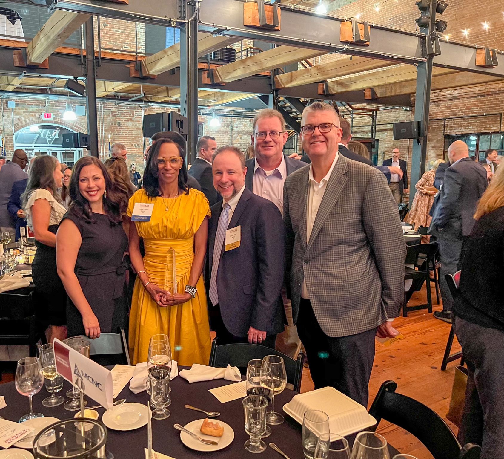 Five people standing together at a social event, smiling, with tables and other guests in the background.