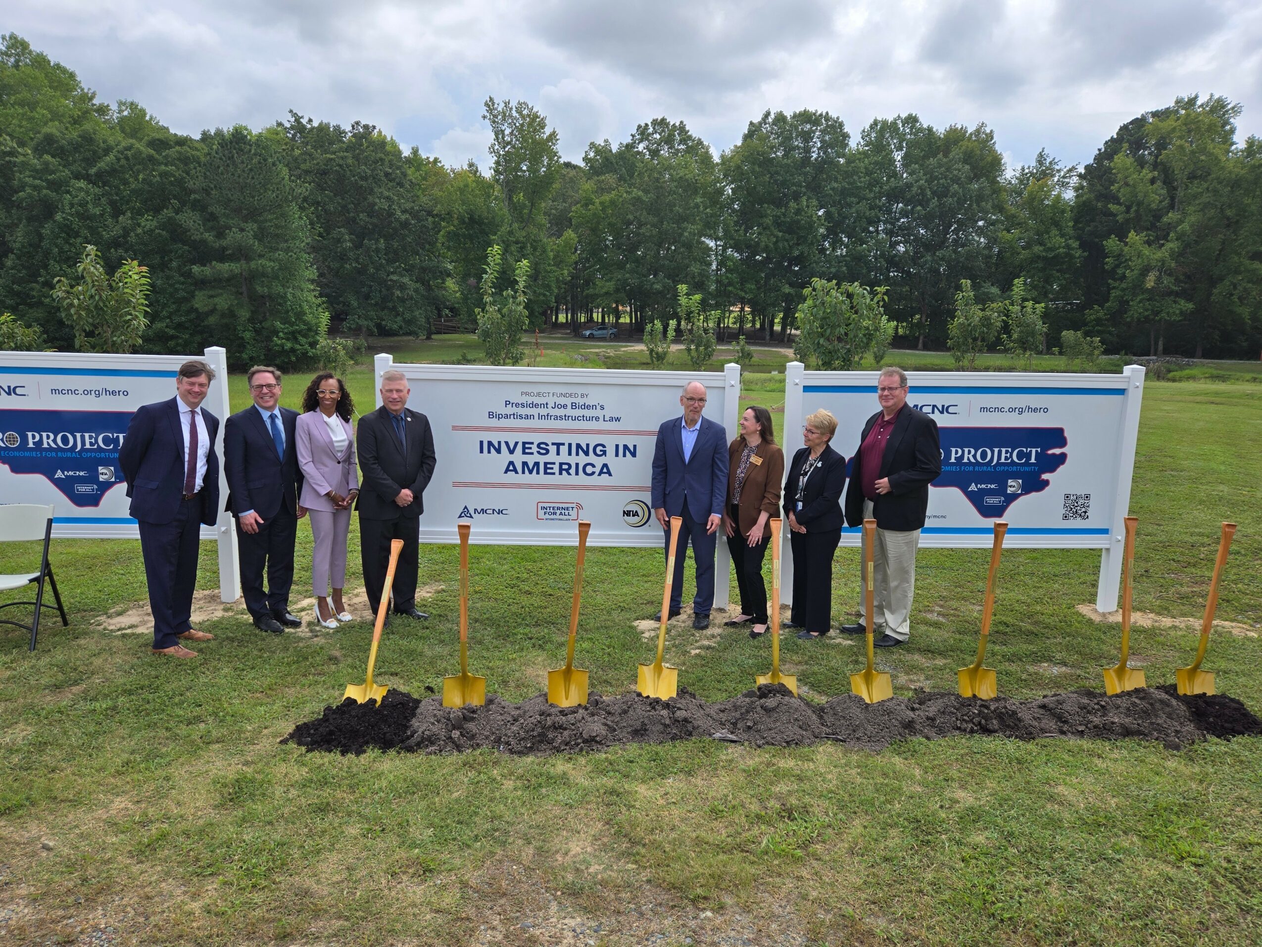 Group of eight people standing outdoors with shovels in front of signs, participating in a groundbreaking ceremony, trees and cloudy sky in background.