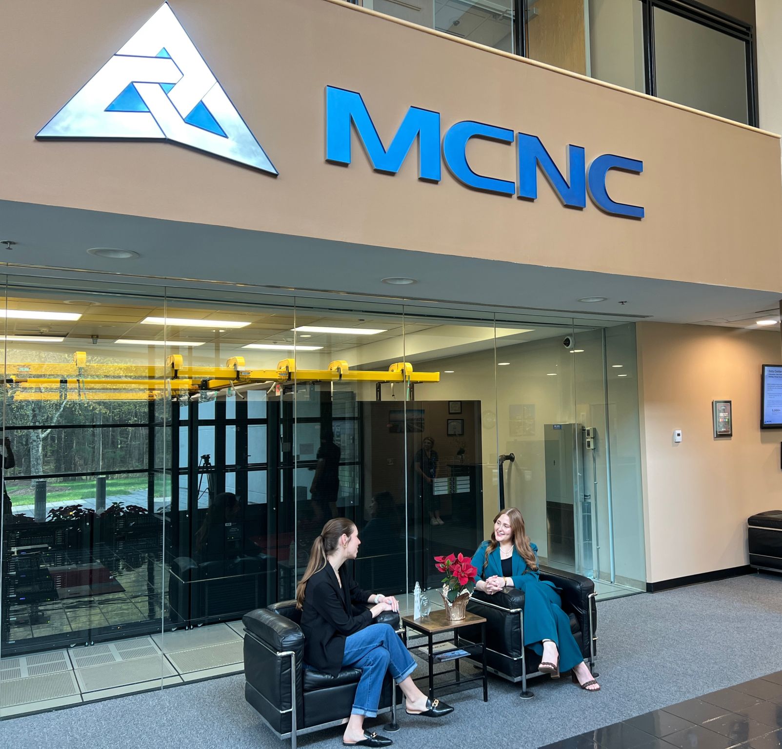 Two women sit and talk outside a building with a large sign reading MCNC, glass doors, and a reception area inside.