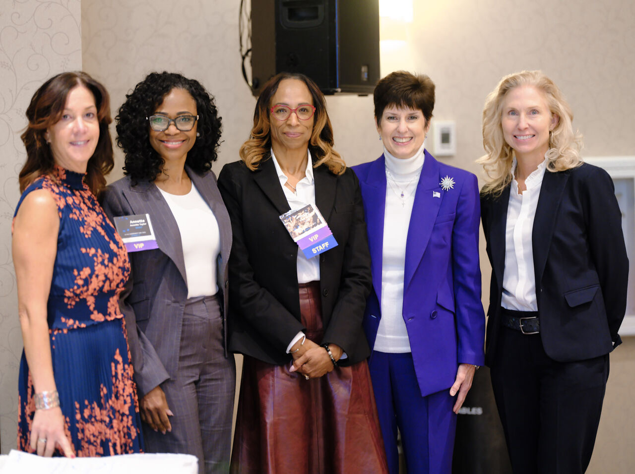 Five women standing together indoors, smiling, dressed in professional attire, with a speaker and wall in the background.