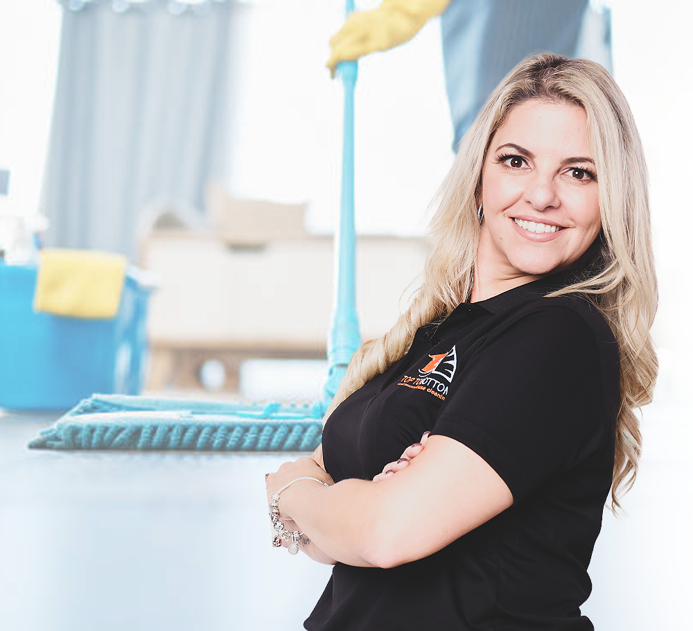Woman smiling with arms crossed, cleaning supplies in background, in a bright room.