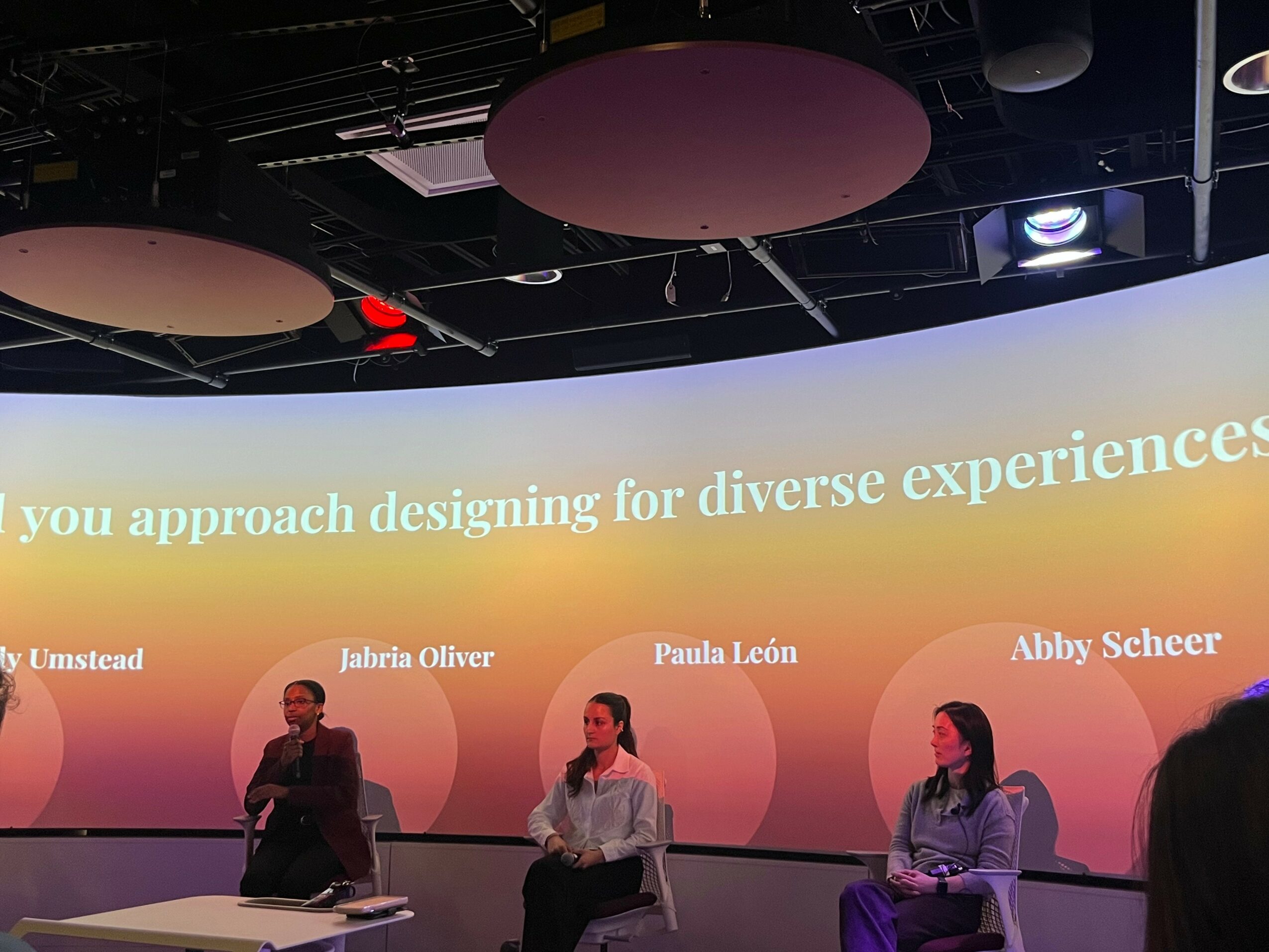 Four people sitting on stage in front of a large screen with text about designing for diverse experiences.