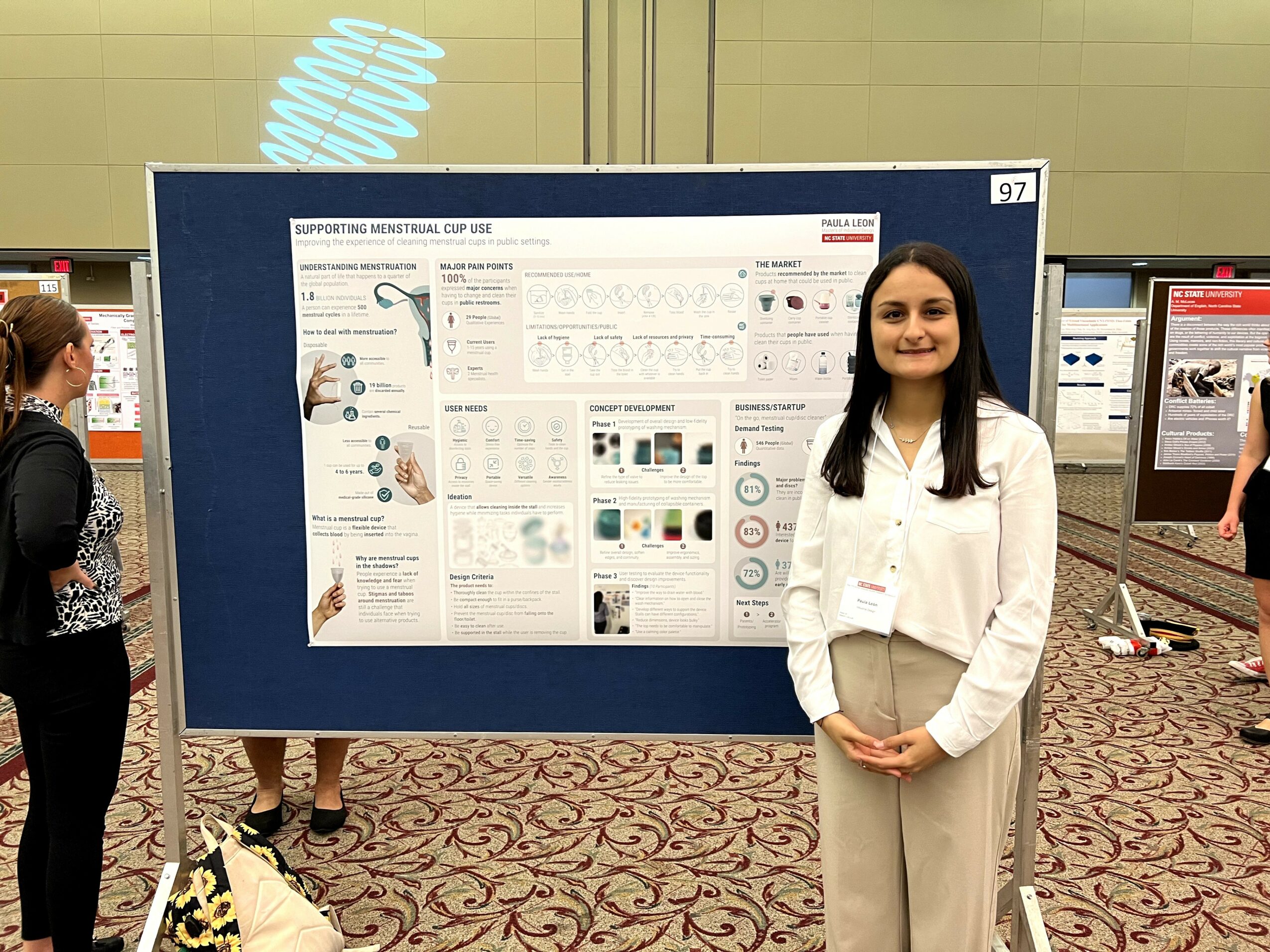 Young woman standing in front of a research poster at a conference, smiling, with other attendees nearby.