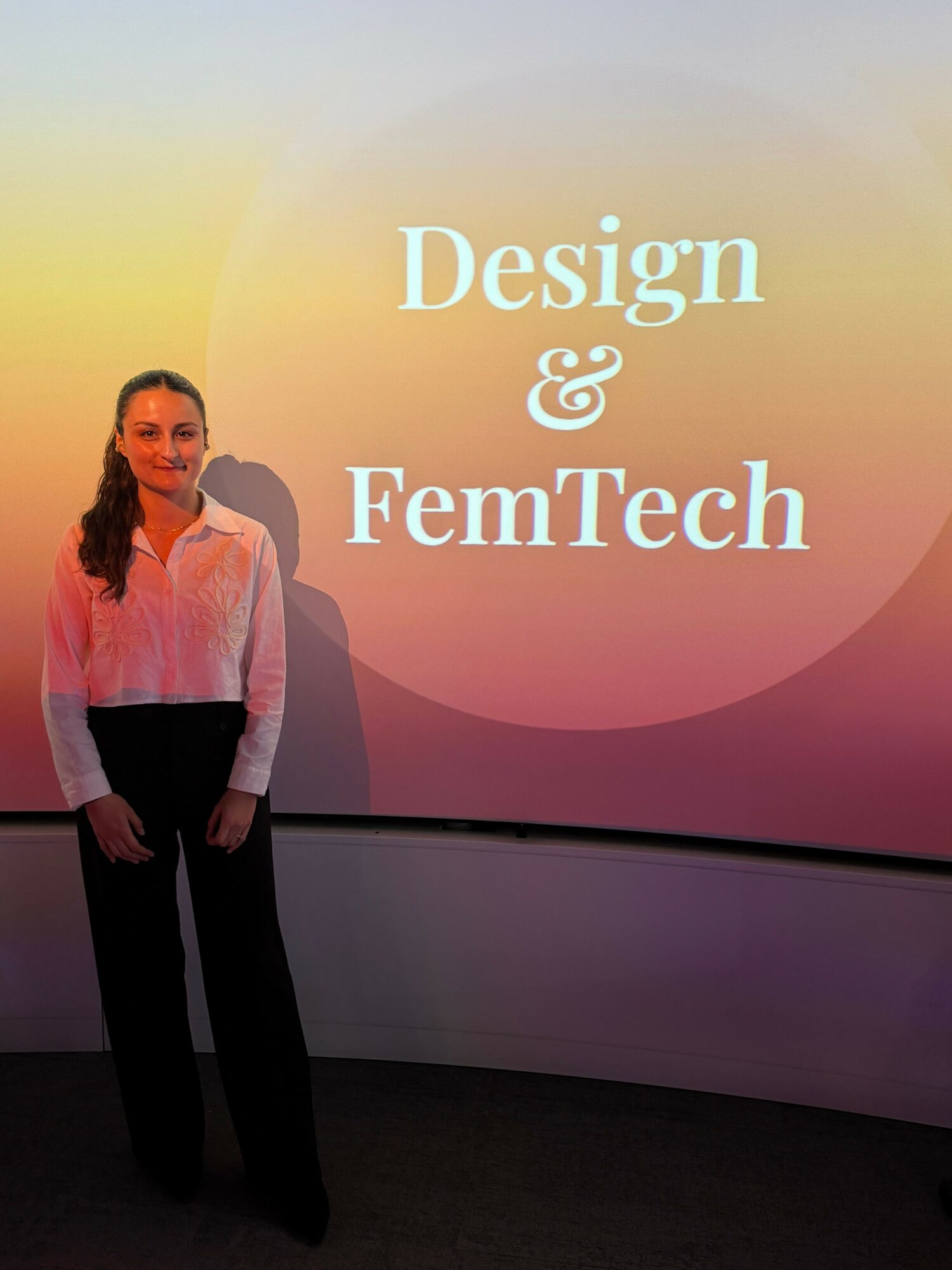 Person standing in front of a colorful background with text about design and FemTech, wearing a light-colored top and dark pants.