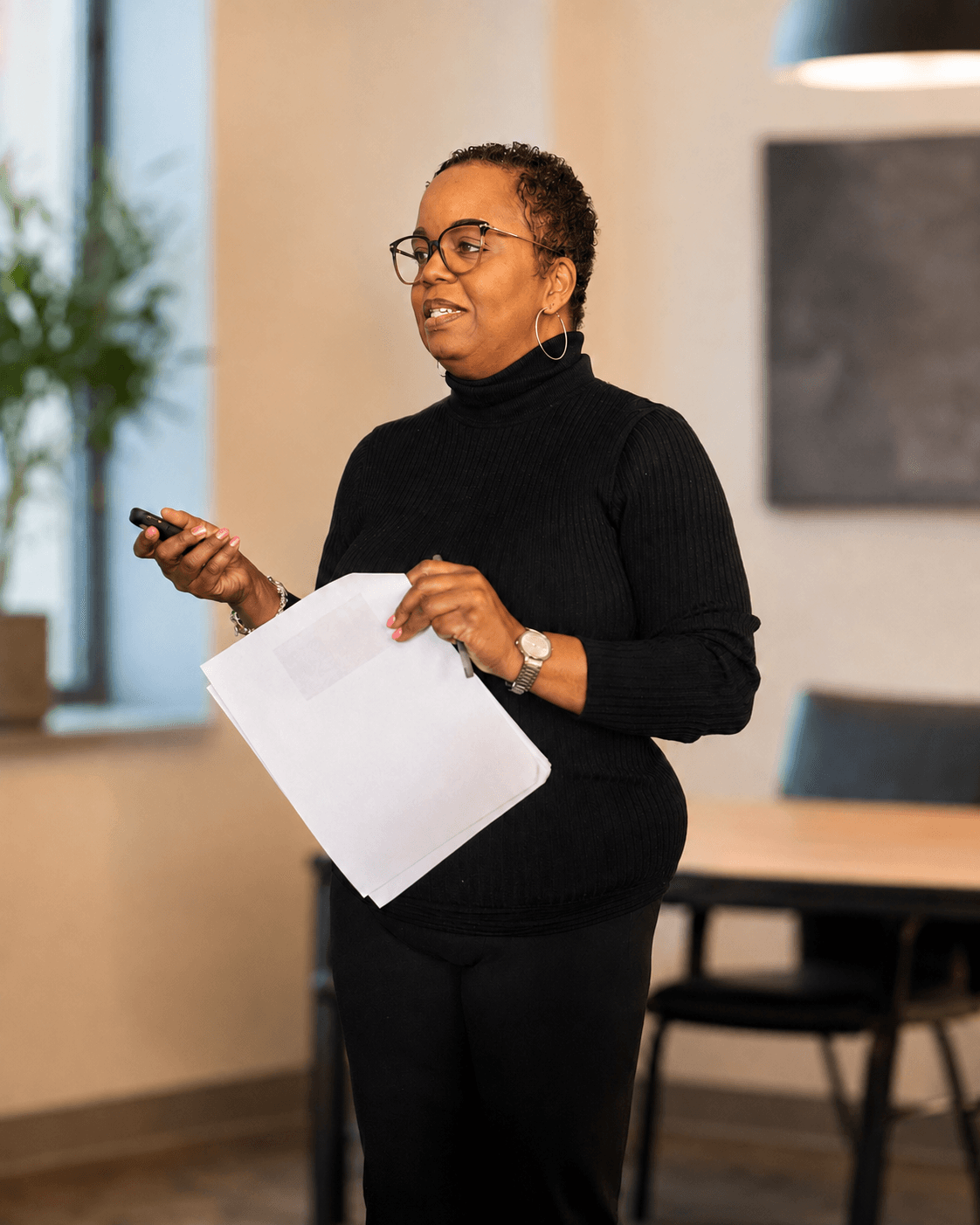 Woman with glasses holding a paper and remote, standing in a room with a table and window.