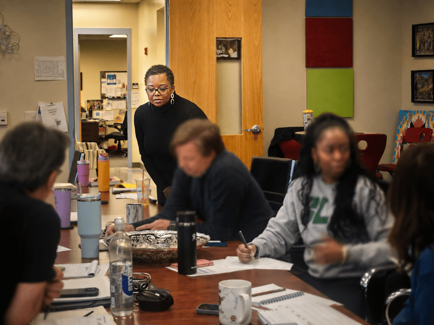 Group of people in a meeting room with a woman standing and others seated around a table, some taking notes.