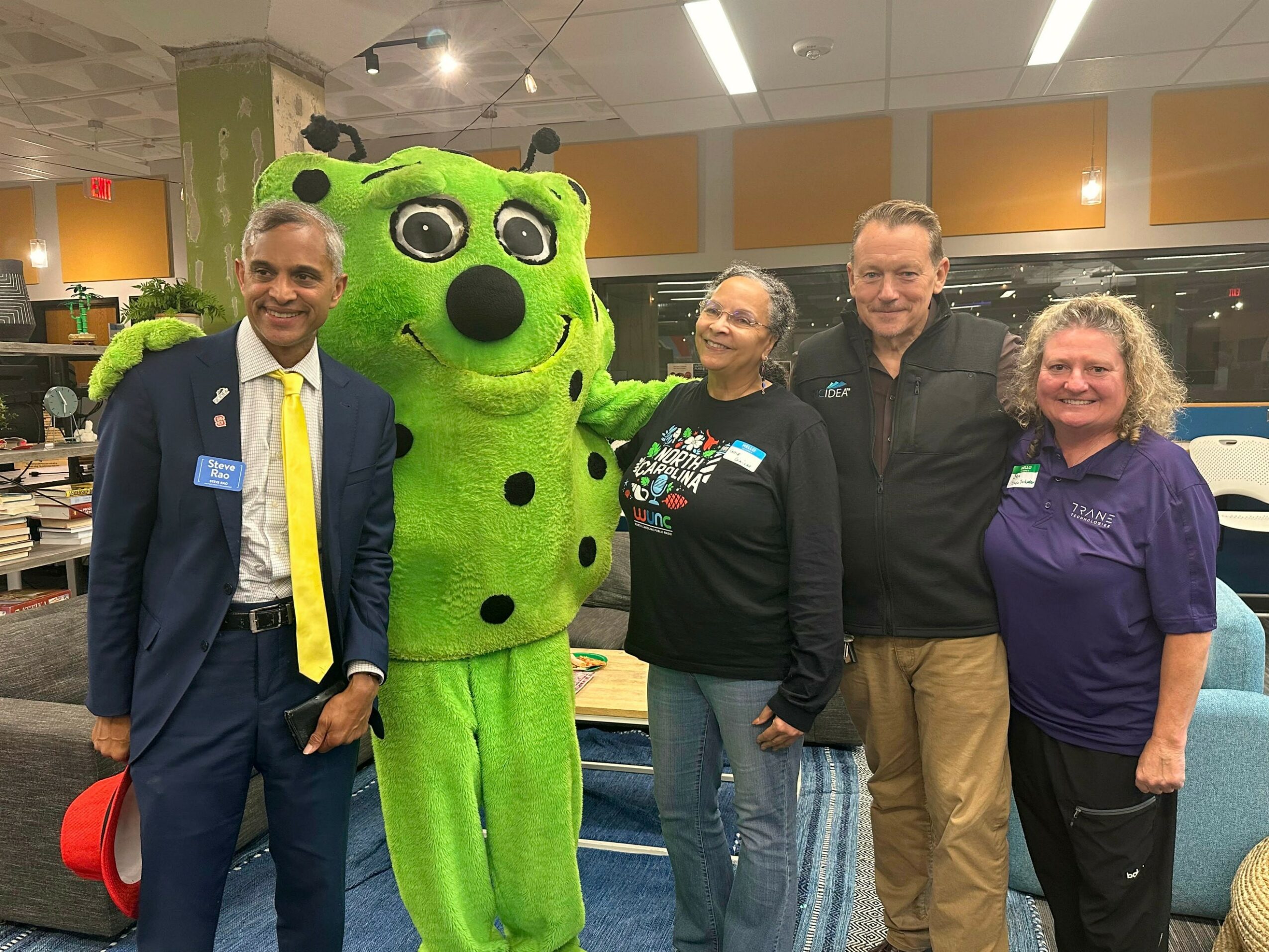 Four people and a green mascot pose indoors, smiling, with tables and chairs in the background.