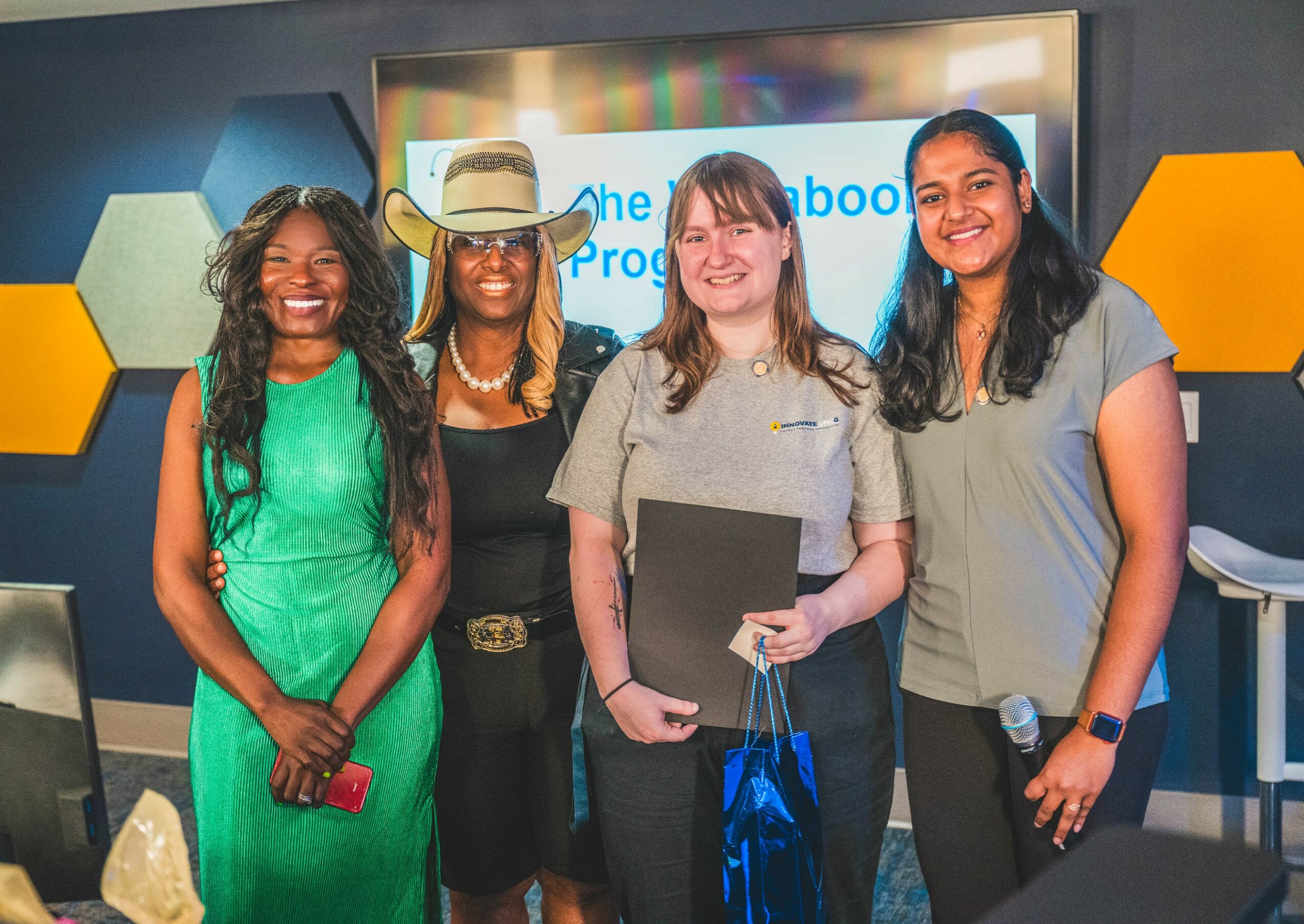 Four women standing together in a room with a screen behind them, smiling and holding items, in a professional setting.