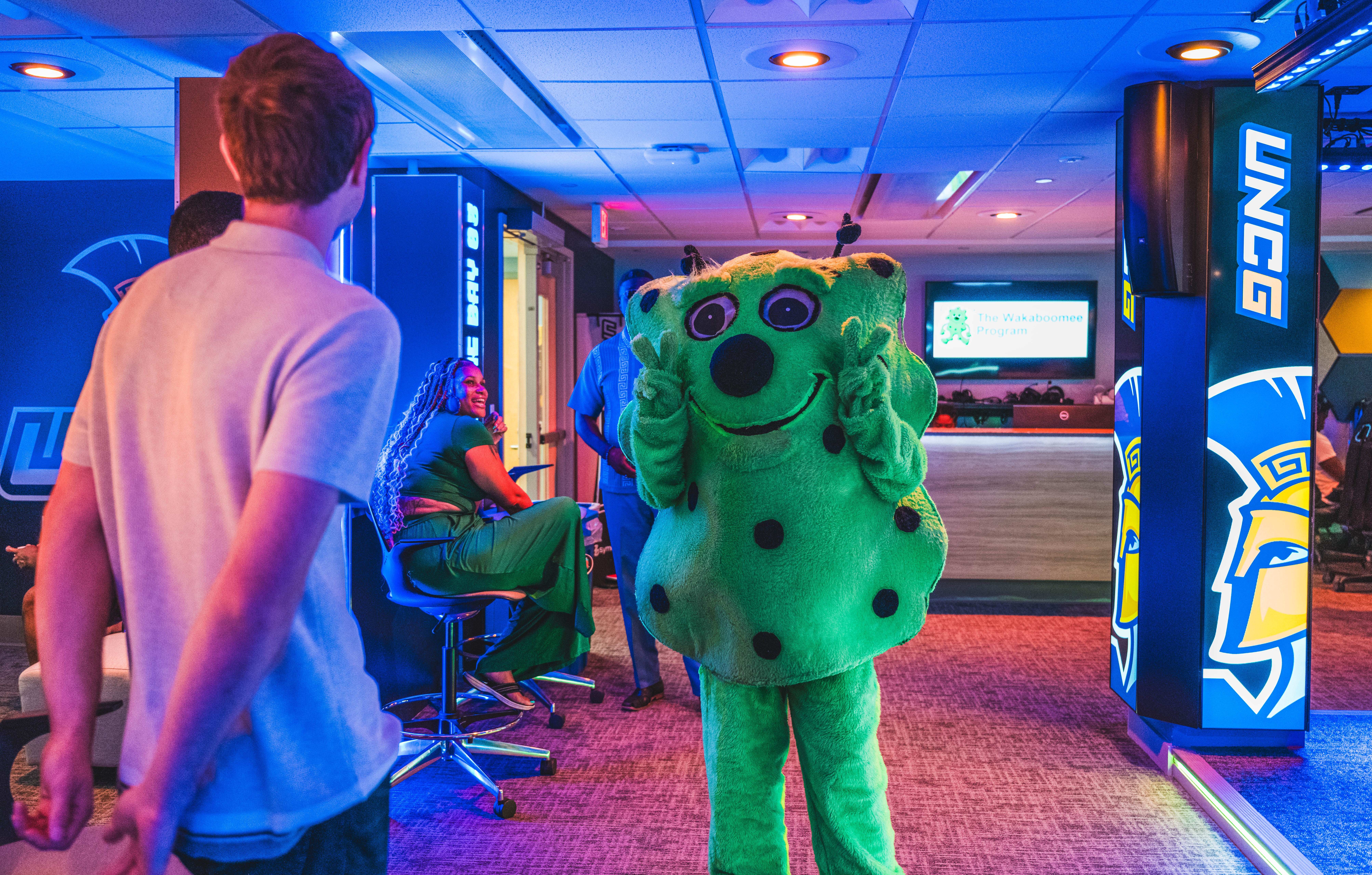 Person in a green mascot costume with black spots and long green hair, in a gaming arcade with screens and neon lights.