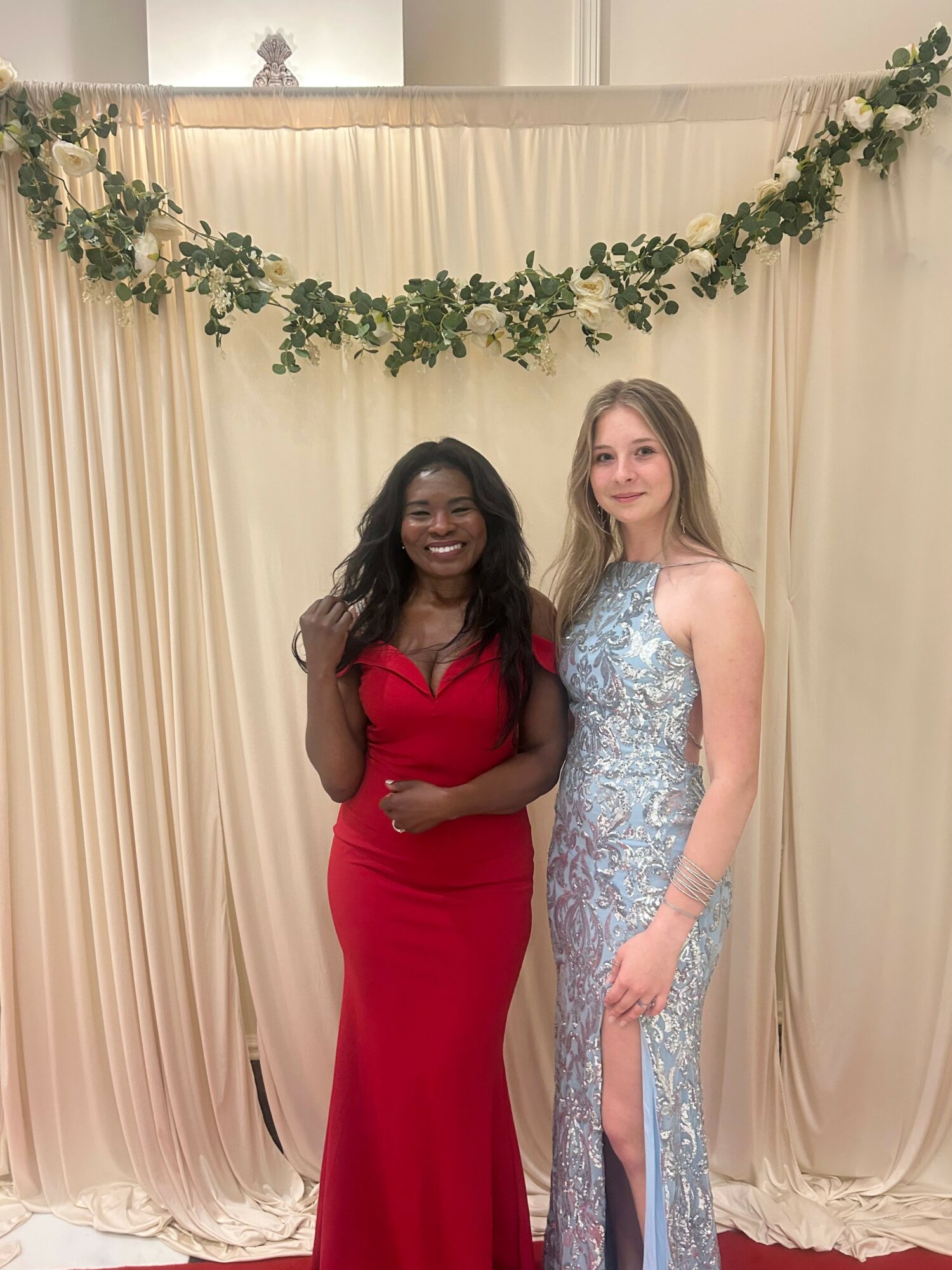 Two women in formal dresses standing in front of a cream-colored curtain with green foliage decoration above, smiling.