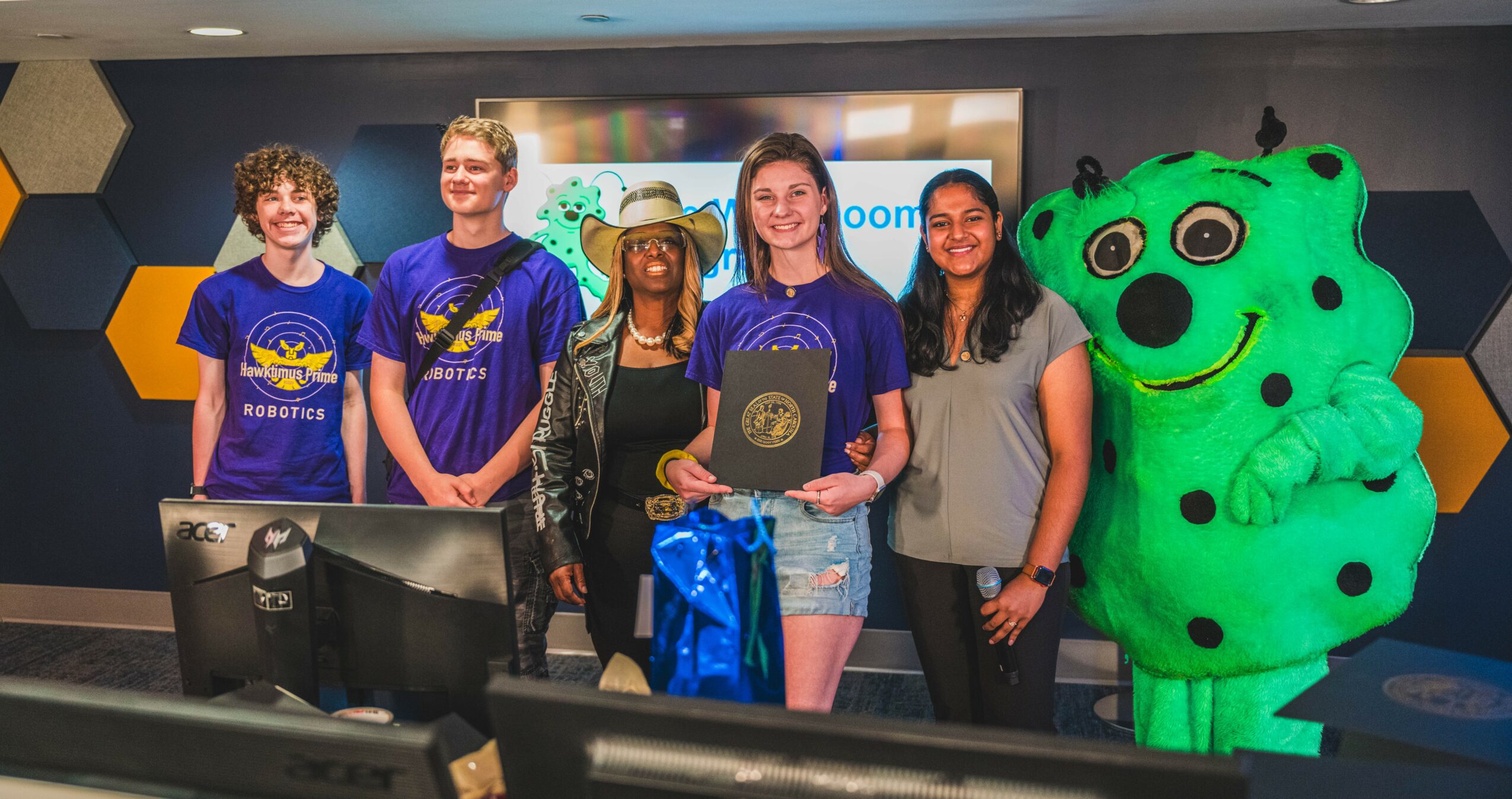 Group of five people and a green mascot mascot standing in front of a screen, some holding awards and gifts.