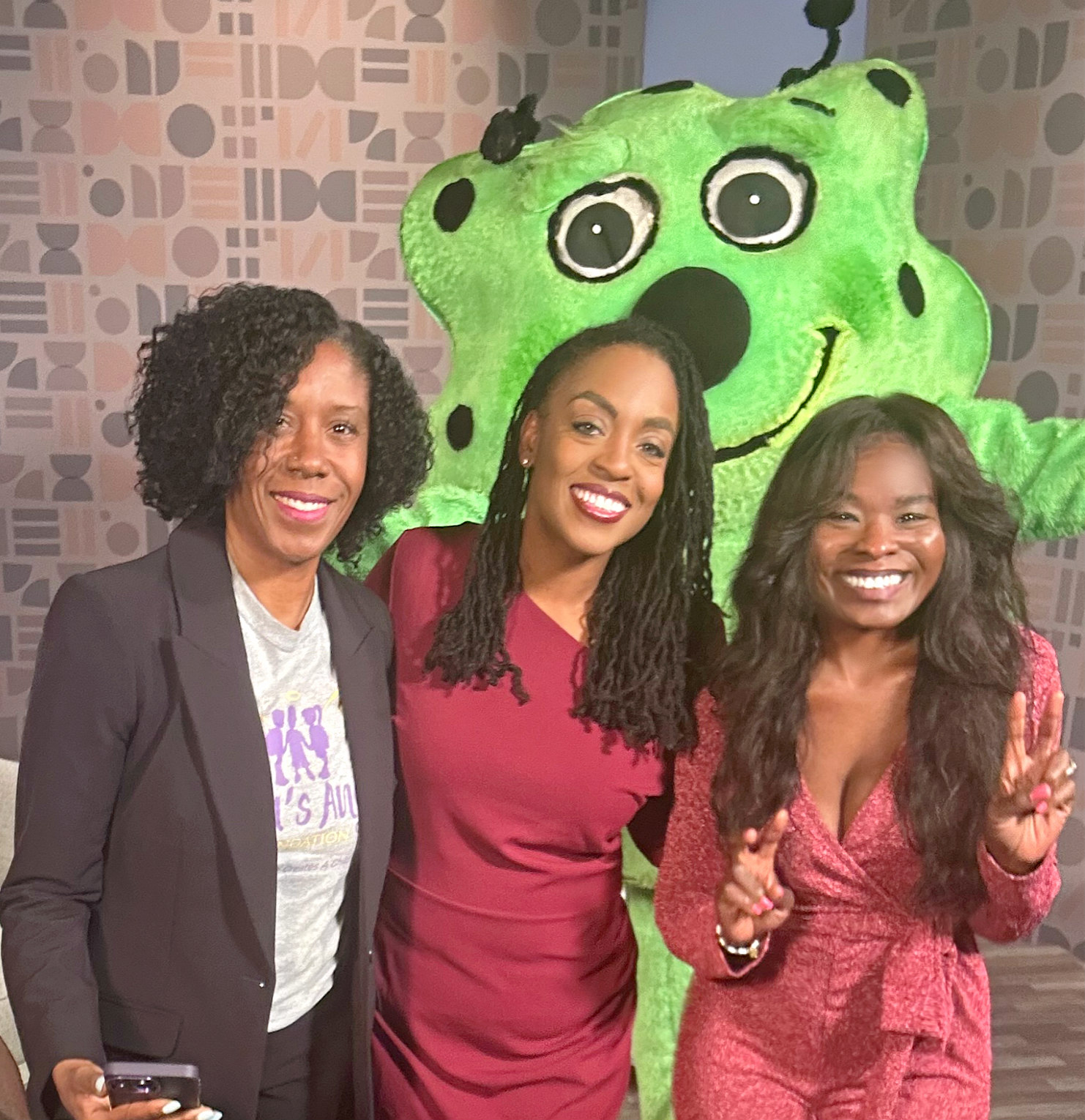 Three women smiling and posing with a green bear mascot in a patterned background.